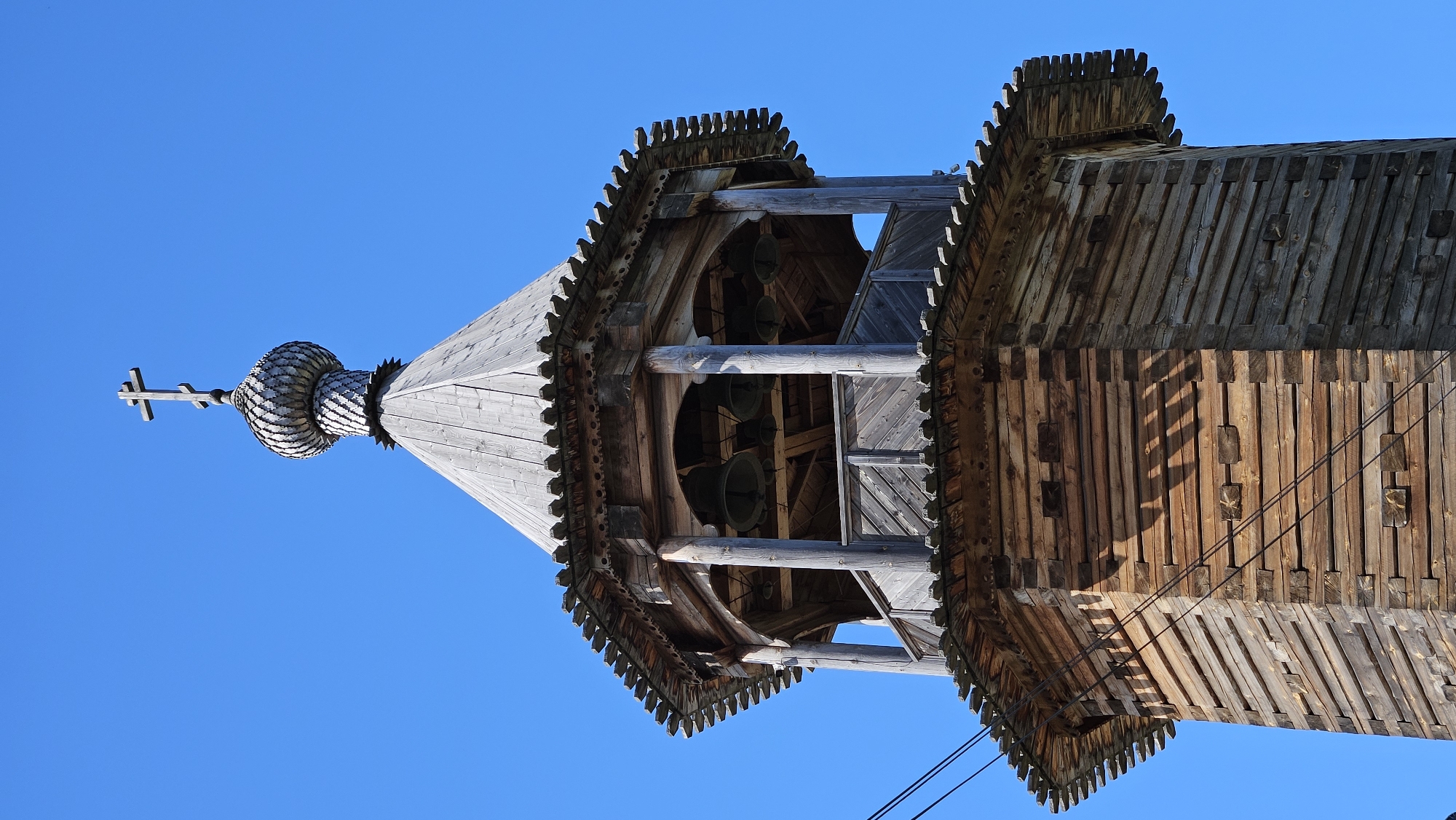 A tall, octagonal wooden bell tower stands against a clear, bright blue sky in Unknown, Unknown. The tower is constructed from horizontal logs with visible interlocking corner joints, exhibiting weathering and variations in wood tone. The upper portion of this log structure is capped by a decorative frieze featuring a sawtooth-like pattern. Above this, an open belfry section is visible, supported by wooden columns forming arched openings. Inside the belfry, several dark metal bells are suspended. A wooden railing encircles the base of the belfry. Above the belfry, another decorative frieze identical to the lower one precedes a steeply pitched, conical wooden roof made of planks. The roof culminates in an onion-shaped dome covered with intricately carved or shingled wooden scales, topped by a three-bar Orthodox Christian cross. Two dark wires traverse the lower left section of the tower diagonally. Sunlight illuminates the structure from the left, casting distinct shadows on the right side of the wooden surfaces. No individuals are present.