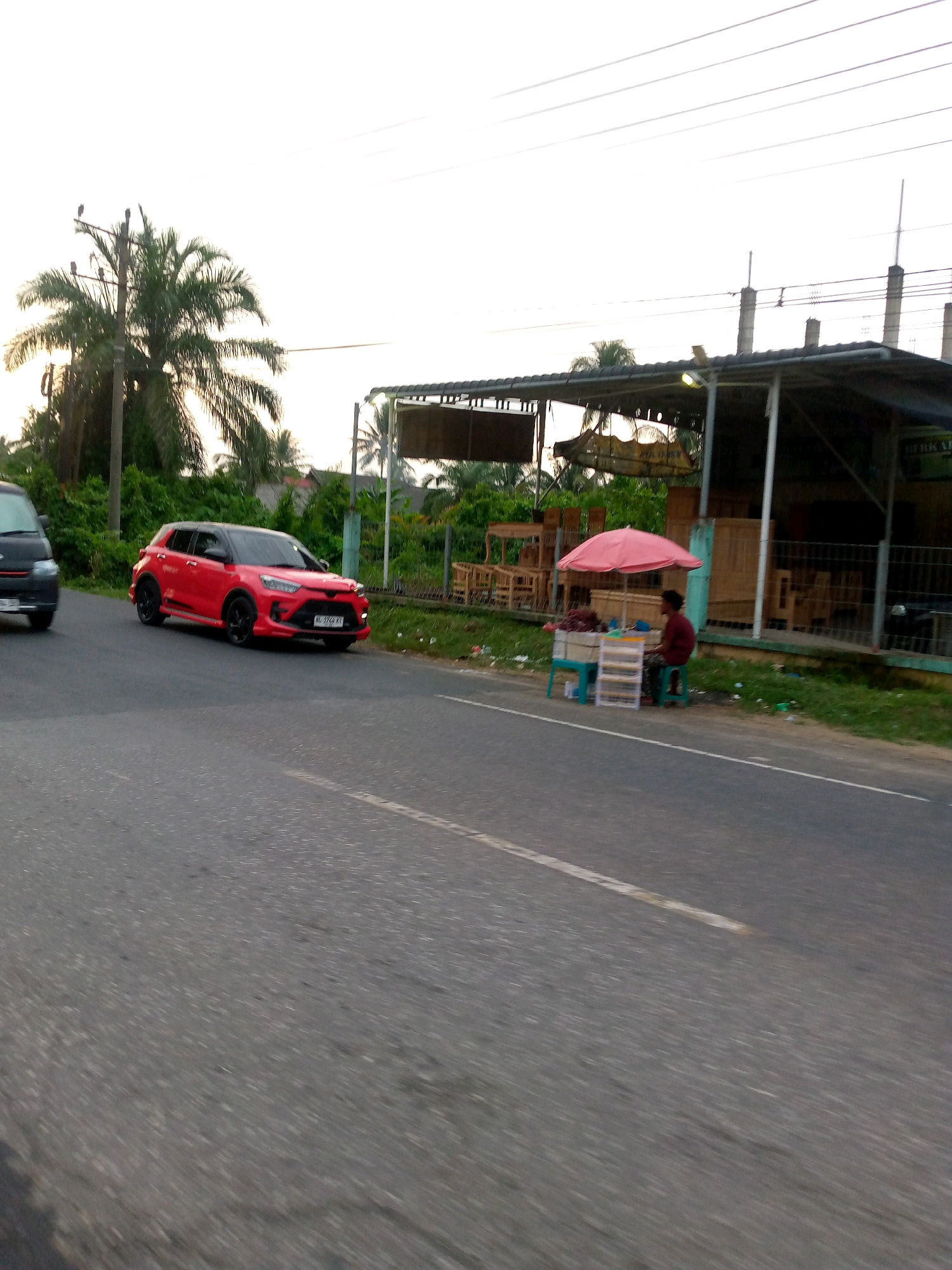 The image depicts a roadside scene in Keudeteupinpunti, Indonesia. A bright red SUV is captured in motion on the left side of the frame, with a dark-colored van partially visible behind it. To the right, a small roadside stall operates under a pink umbrella, offering goods from a display unit. A person in a maroon shirt sits on a stool at the stall. Behind the stall, a building with an open-front structure displays wooden furniture. Palm trees and lush green vegetation border the roadside, and utility poles with overhead wires are present. The sky is overcast and bright. The road surface is asphalt, with a white lane marker visible.
