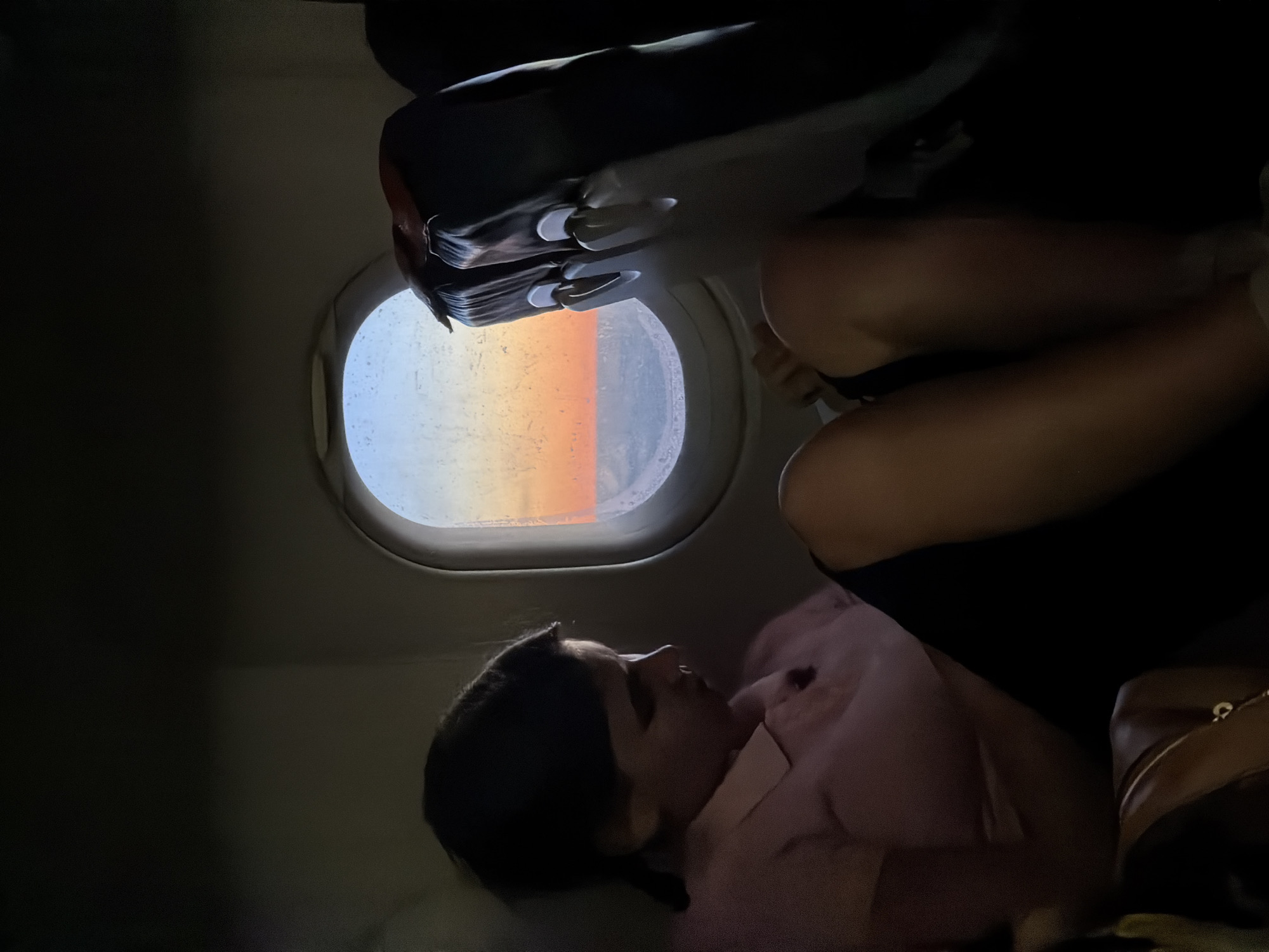 A woman is seen resting on a plane in front of a window that shows a sunset. The setting appears to be on an airplane due to the presence of an airplane window and an overhead console. The image does not show the city of Bobrovka, Russia.
