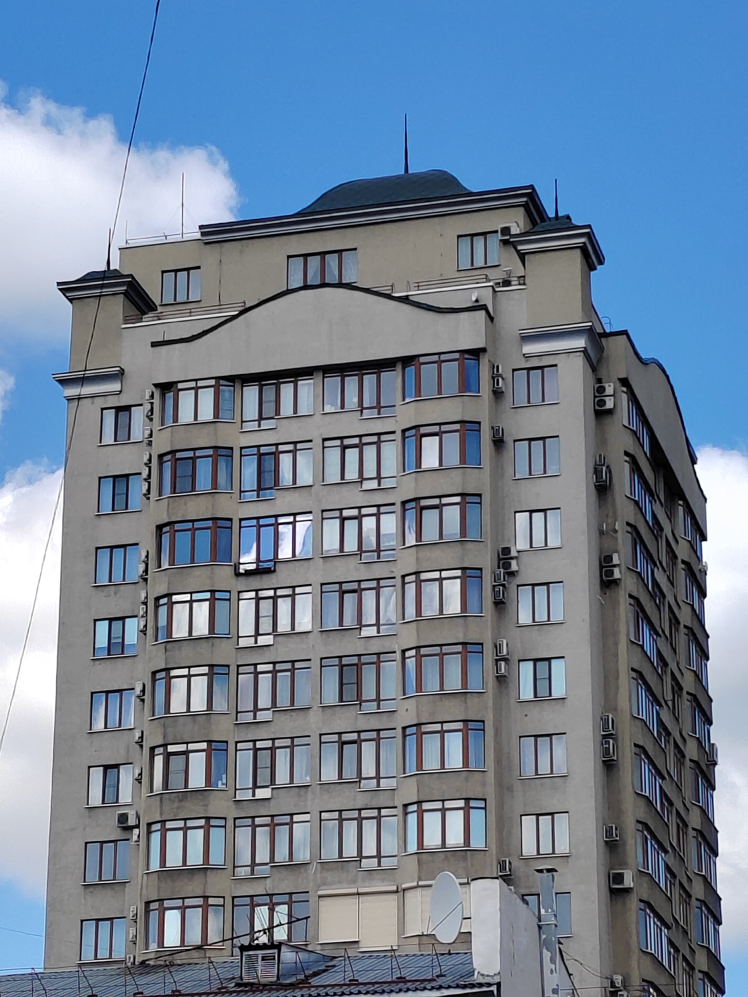 The image shows a tall, modern apartment building in Kemerovo, Russia, against a bright blue sky with scattered white clouds. The building is primarily grey with numerous windows framed in dark brown. Many of the windows appear to be bay windows, giving the building a distinctive, multifaceted facade. Air conditioning units are visible mounted on the exterior walls at various levels. The top of the building features a dark, curved roofline with decorative elements and antennas. The overall impression is that of a residential building in an urban setting.