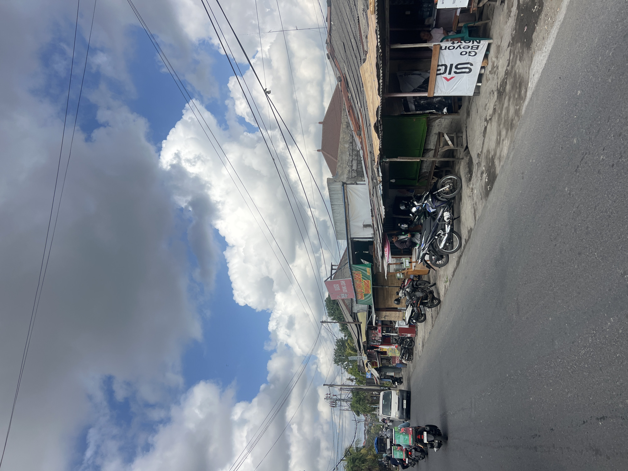 The image depicts a street scene in Denpasar, Indonesia, under a partially cloudy sky. The foreground shows a gray asphalt road with several motorcycles, some carrying delivery boxes, moving along the street. On the right side of the road, a row of modest shops with corrugated metal roofs and open fronts are visible. These shops appear to sell various goods, with some signage in Indonesian. One shop prominently displays a white banner with the text "SIG Go Beyond Next". People are visible within the shopfronts, some standing and others sitting on benches. Further down the street, more vehicles, including a white van and a truck, are present. The background reveals buildings with tiled roofs and some vegetation, suggesting an urban or suburban environment. The overall impression is one of a bustling street with local commerce and everyday activity.