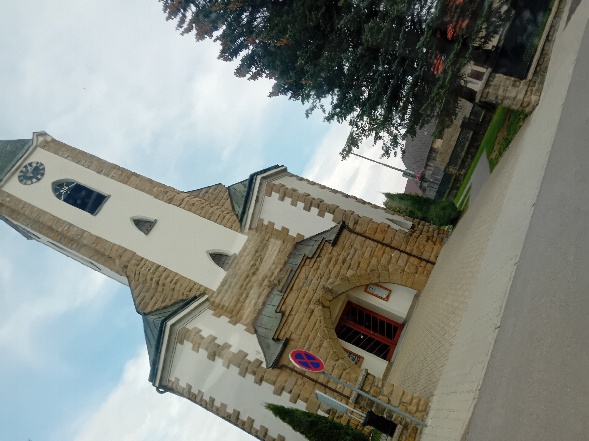 The image shows a tilted view of a church in Zuberec, Slovakia. The church has a white facade with stone accents and a tall tower with a clock. Red double doors lead into the church through an arched stone entrance. A no-parking sign is visible to the left of the entrance. A tall evergreen tree is situated to the right of the church. The sky is partly cloudy, and the overall lighting suggests it is daytime. The foreground shows a paved walkway and a road.