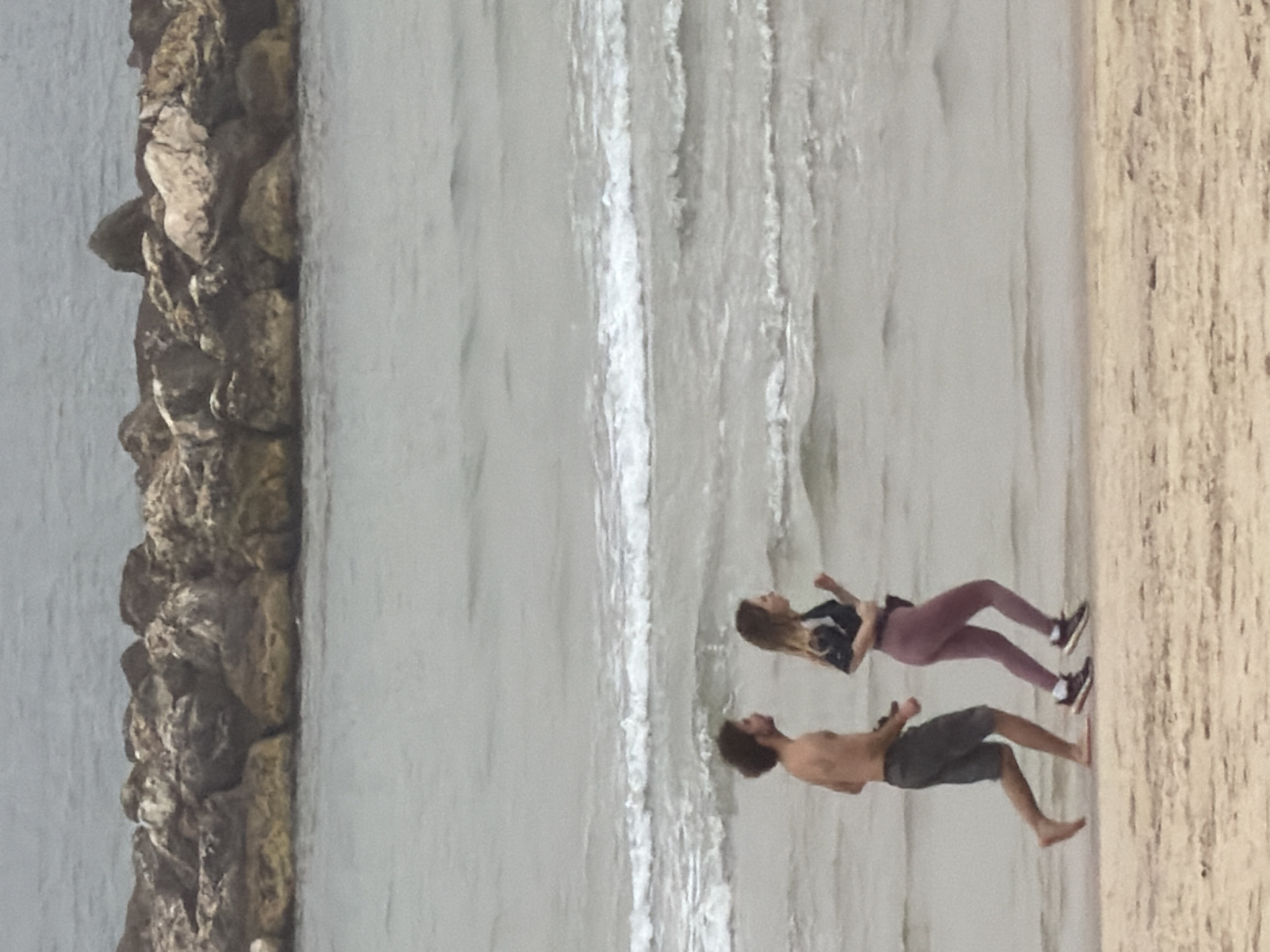 The image shows a man and a woman running along the beach in Tel Aviv, Israel. They are barefoot and close to the water's edge. The man is shirtless and wearing shorts, while the woman is wearing a long-sleeved shirt, leggings, and sneakers. To the left, there is a rock barrier with the sea on the other side. The sand is a light color and the water is mostly white.