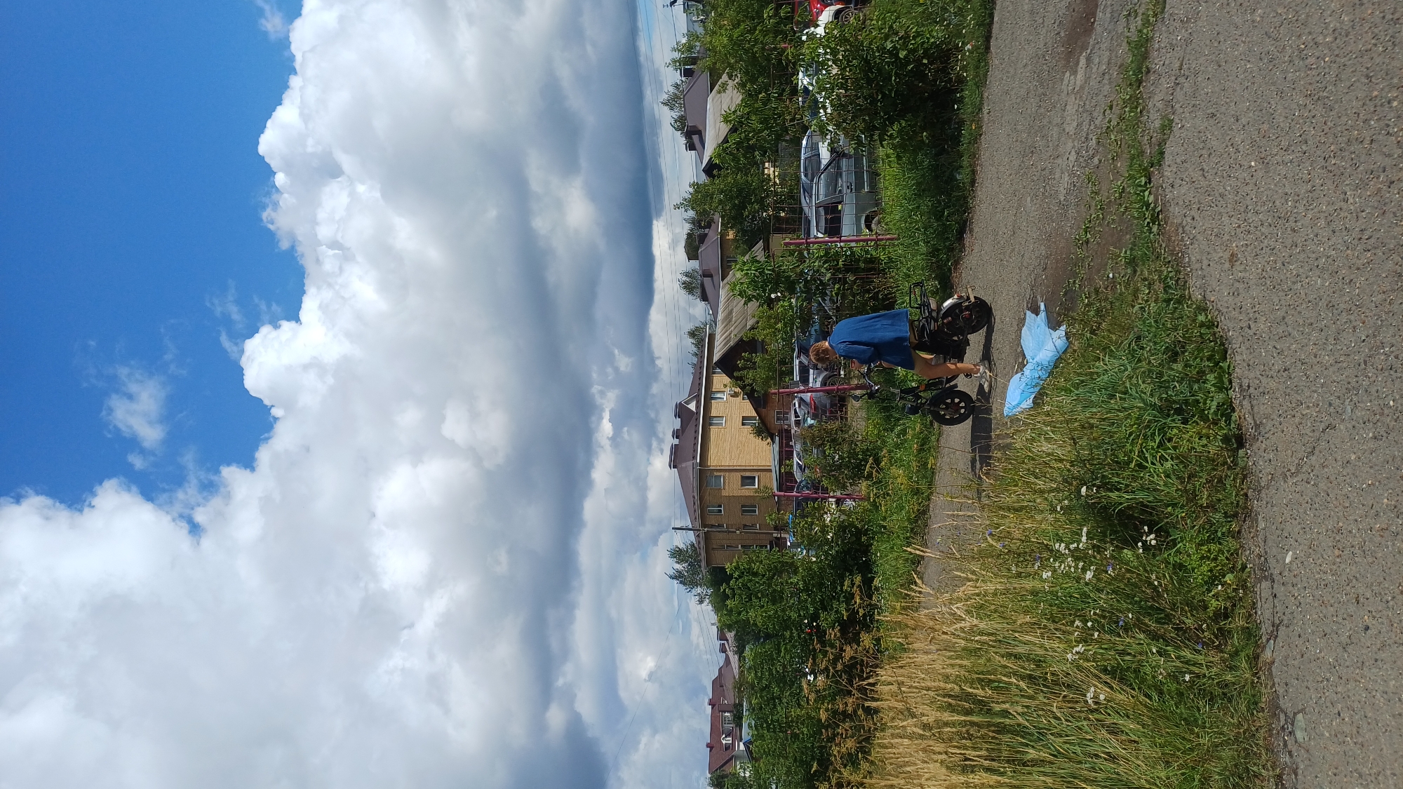 In Izhevsk, Russia, on a paved road surrounded by greenery and vegetation, a person is seen riding a dark-colored electric scooter. The individual, wearing a blue denim jacket and shorts, is positioned on the scooter facing away from the camera. In the background, a light yellow building with a brown roof stands out amidst other houses and trees. A parking lot with several cars is visible behind a purple fence. The sky above is a mix of blue and white clouds, suggesting a partly cloudy day. A crumpled light blue plastic bag lies on the ground near the scooter.