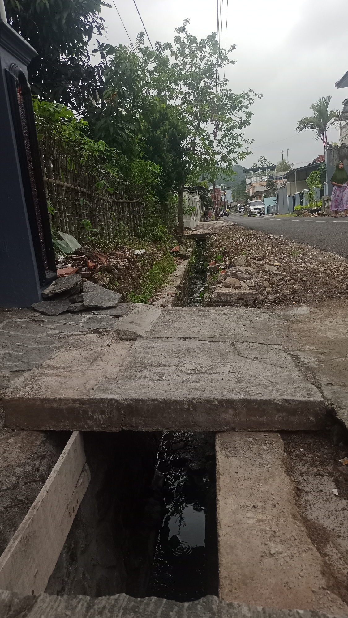 The image depicts a roadside drainage ditch in the city of Pakemitan, Indonesia. In the foreground, a concrete slab covers part of the ditch, with an opening revealing dark water and debris. To the left, a dark blue gate and a fence made of bamboo line the property. Beyond the fence, trees and greenery are visible. The road itself is paved, with a car visible in the distance. To the right of the road, a woman in a green headscarf and a patterned skirt walks away from the camera. The sky is overcast, suggesting a cloudy day. The scene appears to be a typical street view in a residential or semi-urban area.