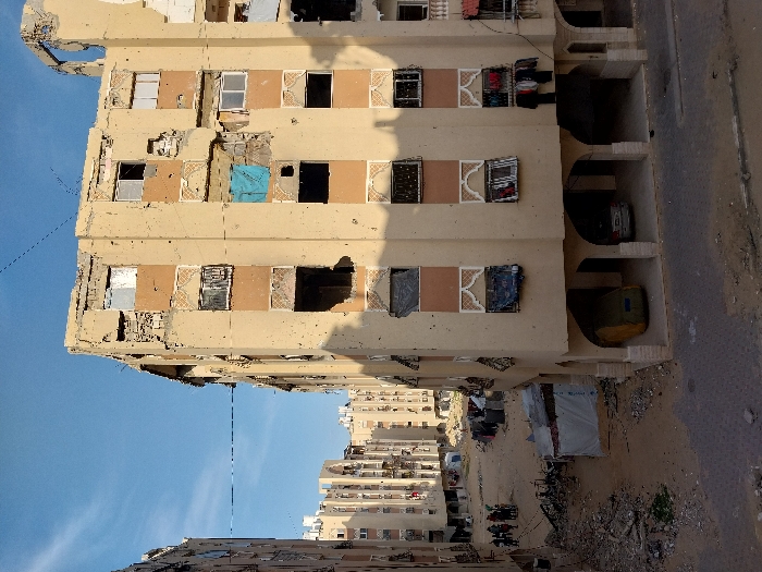 The image presents a severely damaged street scene in Khan Yunis, Palestinian Territory, under a clear blue sky. The prominent subject is a multi-story, light-beige apartment building exhibiting extensive structural damage, including large holes in its facade, shattered window frames, and numerous pockmarks from impacts across all visible floors. Sections of the upper levels show significant material loss and structural compromise. Several windows are either open voids or covered with various fabrics, including a prominent blue tarp on one middle-floor window. Laundry hangs from a line on a lower-level balcony of this building.
The ground level of the main building is supported by arched columns, creating a recessed parking or open area where a beige vehicle covered by a green tarp and a silver car are parked. To the left, a wide, unpaved area of dirt and rubble extends between the central building and another similar, also damaged, multi-story apartment building. Within this rubble-strewn space, several makeshift shelters constructed from tarps are visible, one prominently displaying the UNICEF logo. A group of approximately five to seven individuals is gathered near these shelters on the dirt ground. Further in the background, more apartment buildings, similarly affected, line what appears to be an unpaved urban thoroughfare. The immediate foreground consists of a cracked and worn paved road surface transitioning into the dirt and debris.