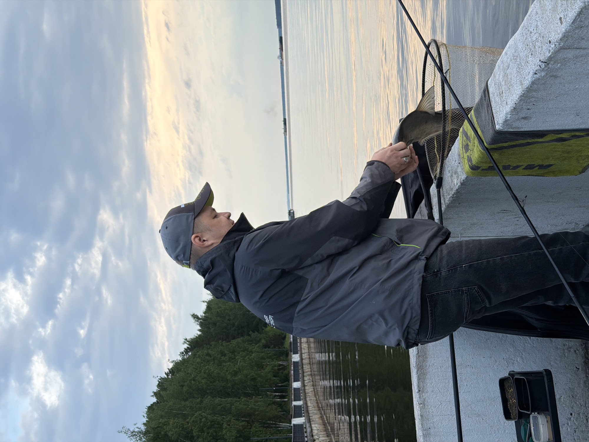 An outdoor image, rotated counter-clockwise, depicts a man fishing from a concrete structure in Chaikovsky, Russia. The man, positioned in profile facing right, wears a grey baseball cap, a dark grey and black waterproof jacket, and dark jeans. He is actively handling a fish, holding it with both hands near a dark-framed landing net with yellow mesh. The fish, appearing medium-sized, is partially visible.
The man stands on a substantial grey concrete pier or embankment. To his lower right, a black tackle box or bait box containing bait and small containers is visible. A fishing rod leans against the concrete structure near his right leg. On the side of the concrete structure, a section is covered with a yellow and black material, possibly for grip or safety.
The background features a vast body of water, likely a river or lake, reflecting the soft light from the sky. Across the water, a distant shoreline is visible, densely covered with green trees. The sky above is partly cloudy, with hues of soft blue and a warm, diffused light suggesting either early morning or late afternoon.
Visible text is limited; on the man's jacket, near his left arm, faint text reading "ENER..." or similar, possibly part of "ENERGO REGION" or "ENERGY REGION," can be discerned. No other clear text is legible on the cap, net, or other objects. The scene conveys a calm moment of recreational fishing.