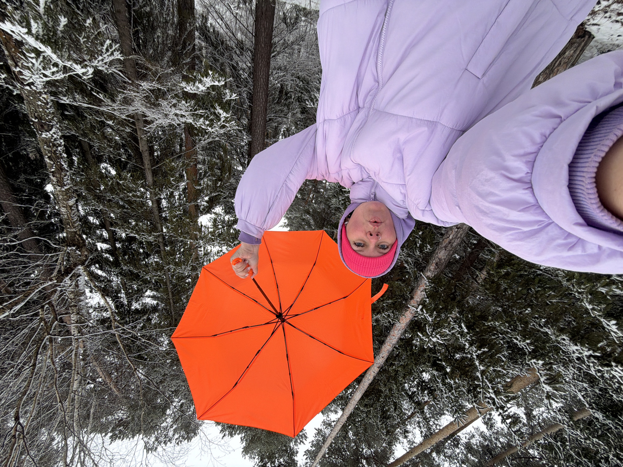 A person wearing a lavender puffy coat and a pink beanie is holding an orange umbrella. The person is positioned against a backdrop of snow-covered trees in a forest, possibly in or near the city of Chaikovsky, Russia. The image appears to have been rotated so the person is upside down in the frame.