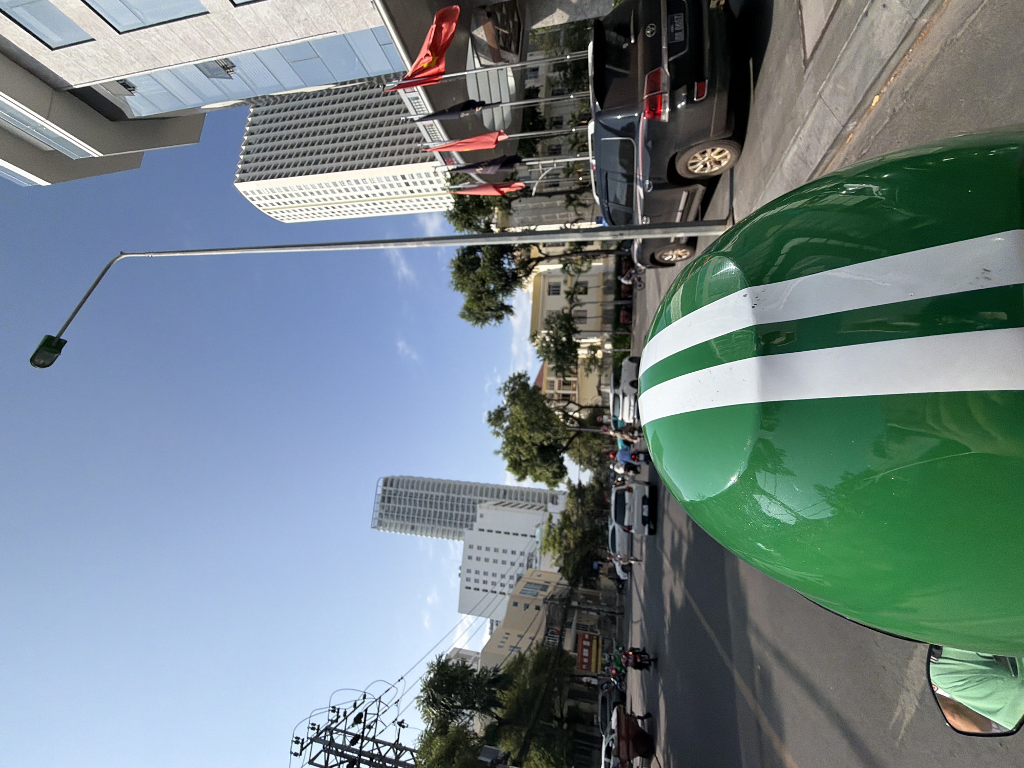 The image is a point-of-view shot from a moving vehicle, likely a motorcycle, in Nha Trang, Vietnam. The foreground is dominated by the green helmet of the rider, with two white stripes. A side mirror reflects a person wearing a green t-shirt. The vehicle is on a paved road with visible lane markings and a curb.
In the middle ground, several cars and motorcycles are in motion on the road. To the right, a black SUV with a license plate reading "BB 0657" is parked. A modern, light-colored building with numerous windows and balconies stands prominently on the right side of the street. Several flags are displayed on poles in front of this building, including what appears to be the Vietnamese national flag.
The background features a mix of urban elements: taller, modern buildings with repetitive window patterns, smaller buildings, trees, and utility poles with many wires. The sky is clear and blue. The overall impression is a bustling city street on a sunny day.