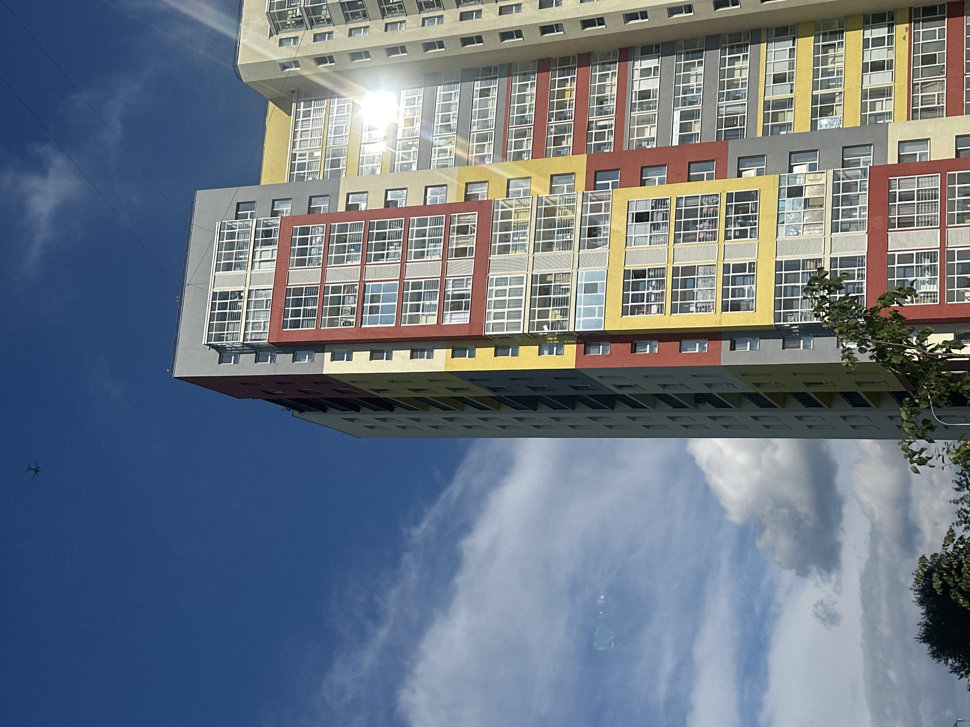 An outdoor, eye-level, vertical shot shows the side of a modern, multi-story residential building with a brightly colored facade of yellow, red, and gray sections. Multiple rows of windows, many with balconies, are visible on each level. The building is illuminated by bright sunlight, creating lens flare in the upper right quadrant of the image. In the distance, a small airplane is visible in the clear blue sky, with some scattered white clouds. The lower portion of the image shows the tops of trees and foliage. This scene is likely from the city of Perm, Russia, where buildings of this architectural style are common.