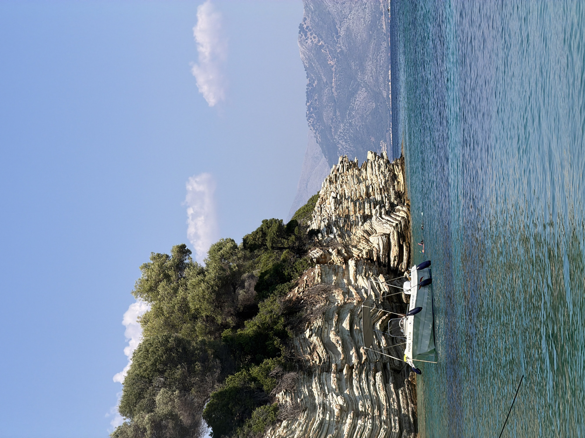 This is an eye-level, long shot photograph of a rocky coastline with lush green trees on top, under a clear blue sky with a few scattered clouds. In the foreground, the turquoise water is calm with subtle ripples. A white boat with a canopy is anchored near the shore, with blue fenders attached. Two individuals are visible in the water near the boat, appearing to be swimming or snorkeling. In the background, hazy mountains rise from the sea. The overall scene depicts a tranquil coastal environment in the Municipal Unit of Kastos, Greece.