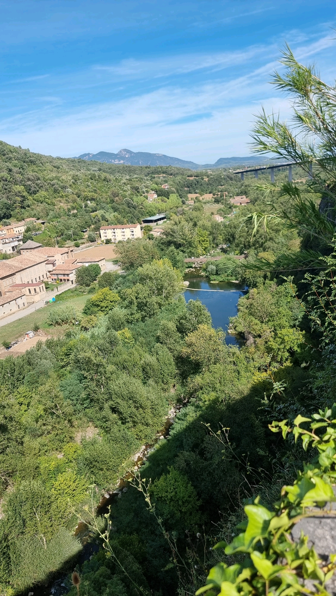 The image depicts a scenic view overlooking the village of Castellfollit de la Roca, Spain. In the foreground, lush green foliage and vegetation dominate the right side of the frame, casting a shadow across the lower portion of the scene. A narrow, rocky stream winds through the dense greenery in the lower part of the image. In the middle ground, a dark blue body of water, likely a river, is visible, crossed by a slender pedestrian bridge. To the left of the river, several buildings with terracotta roofs are clustered together, indicative of a village or town. Behind these structures, a taller, light-colored building with multiple floors stands out. Further in the background, rolling hills covered in dense green trees rise up, leading to distant mountains under a bright blue sky with wispy white clouds. A modern elevated bridge with concrete pillars and a wide deck is a prominent feature in the upper right portion of the background, spanning across the valley. The overall impression is one of a tranquil, verdant landscape with a blend of natural beauty and human settlement.
