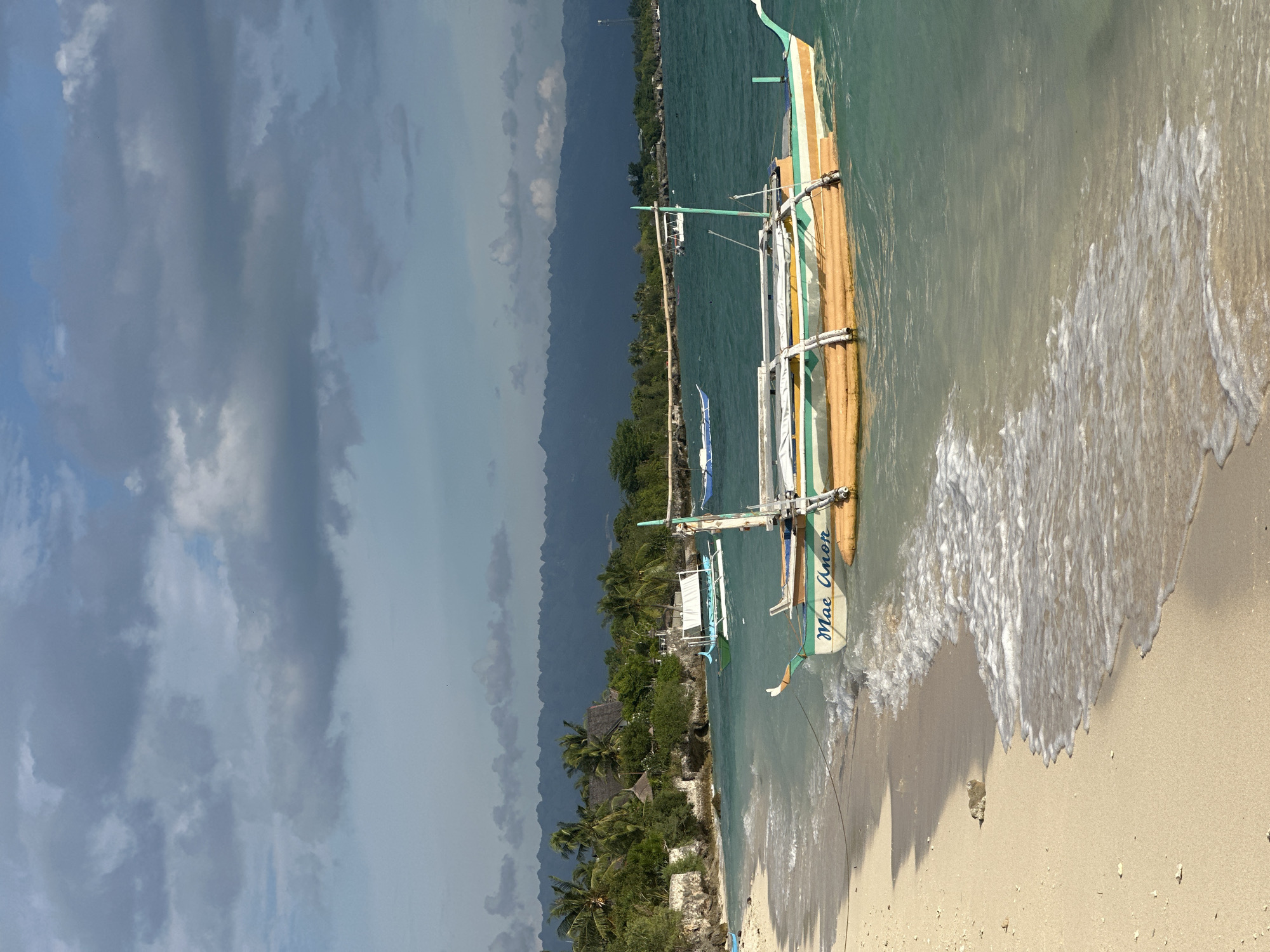 An over-the-shoulder shot shows a beach scene in Saavedra, Philippines. A tan sand beach is in the foreground, with waves gently lapping the shore. A narrow rock protrudes from the sand near the water's edge. A small, outrigger boat named "Mae Anon" is tied to the shore, bobbing in the turquoise water. The boat is painted with orange, green, and white stripes. Beyond the boat, several other boats are visible on the water, some with white awnings. In the distance, lush green trees and palm trees line the shore, with thatched-roof buildings nestled amongst them. A chain of dark, misty mountains forms the horizon, silhouetted against a sky filled with dramatic, grey clouds.