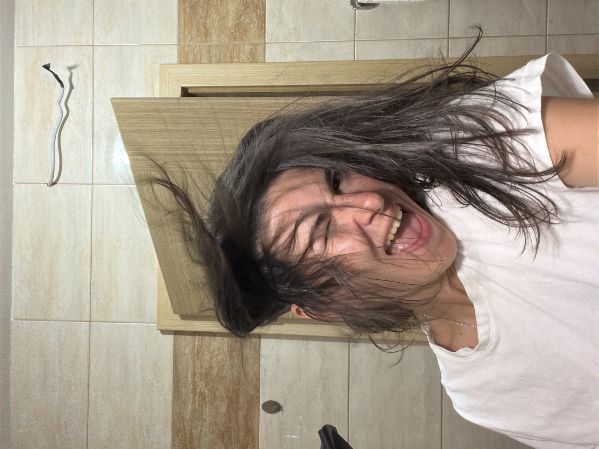 The image depicts a woman with dark hair and a white t-shirt, laughing with her eyes closed. She is positioned in what appears to be a bathroom, with tiled walls in a beige and brown color scheme. A wooden cabinet is visible behind her head. A detached white and black cable hangs on the wall to the left. A small, dark object is on the floor. The location is possibly in Budva, Montenegro.
