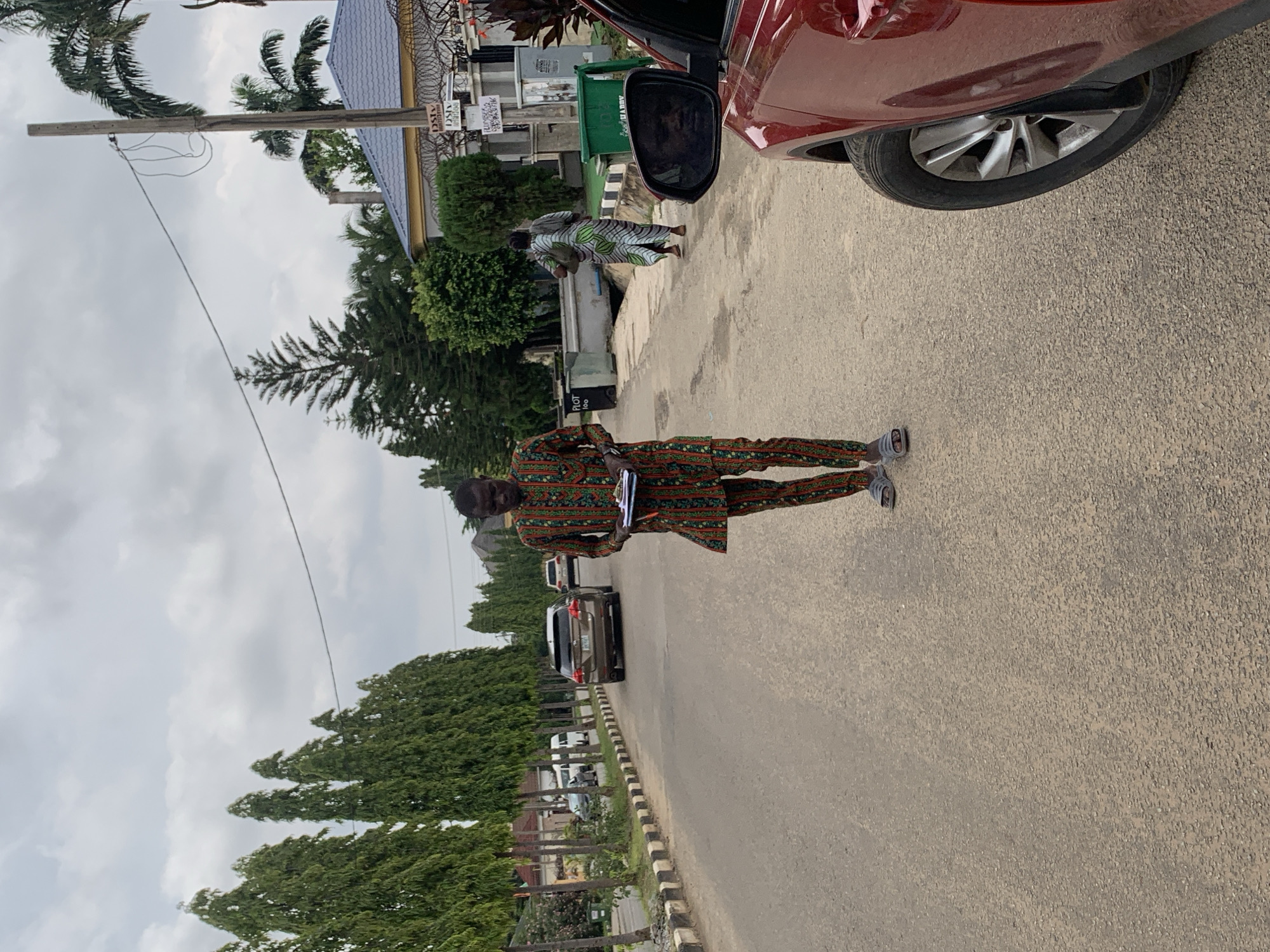A man in a colorful patterned outfit walks down a paved road in Abuja, Nigeria, holding a clipboard and pen. He is wearing light-colored sandals. To his right, the side mirror of a red vehicle reflects his face. In the background, a woman in a patterned dress walks away from the camera, carrying a bag over her shoulder. Trees line the left side of the road, and several cars are parked or driving on the street. Buildings and fences are visible in the distance. The sky is overcast.