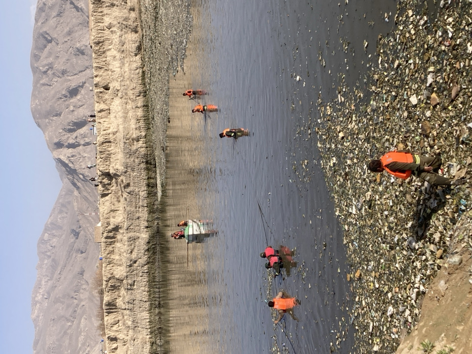 This image depicts a scene in Quetta City Tehsil, Pakistan. A wide, shallow body of murky water, heavily polluted with floating refuse, occupies the central and foreground areas. The immediate foreground bank is a sloping dirt embankment, densely covered with trash including plastics, paper, and other debris.
In the foreground, one individual, wearing an orange life jacket and dark trousers, is seen crouching or kneeling on the trash-laden bank, facing the water. In the midground water, five individuals are present, all wearing orange or red life jackets. Three are wading, with one actively pulling a dark rope or line through the water. Two additional individuals are visible further back in the water. A small, light-colored boat, containing two persons also wearing life jackets, floats in the midground; one person in the boat holds an oar. The water's surface is significantly obscured by a widespread accumulation of floating garbage.
The far bank rises as a steep, eroded earthen cliff. Several small figures are visible walking or standing along the top of this cliff. Beyond the far bank, a range of barren, light-brown mountains extends across the horizon under a clear, bright sky. The individuals in the water and boat appear to be engaged in a clean-up or retrieval operation within the polluted waterway.