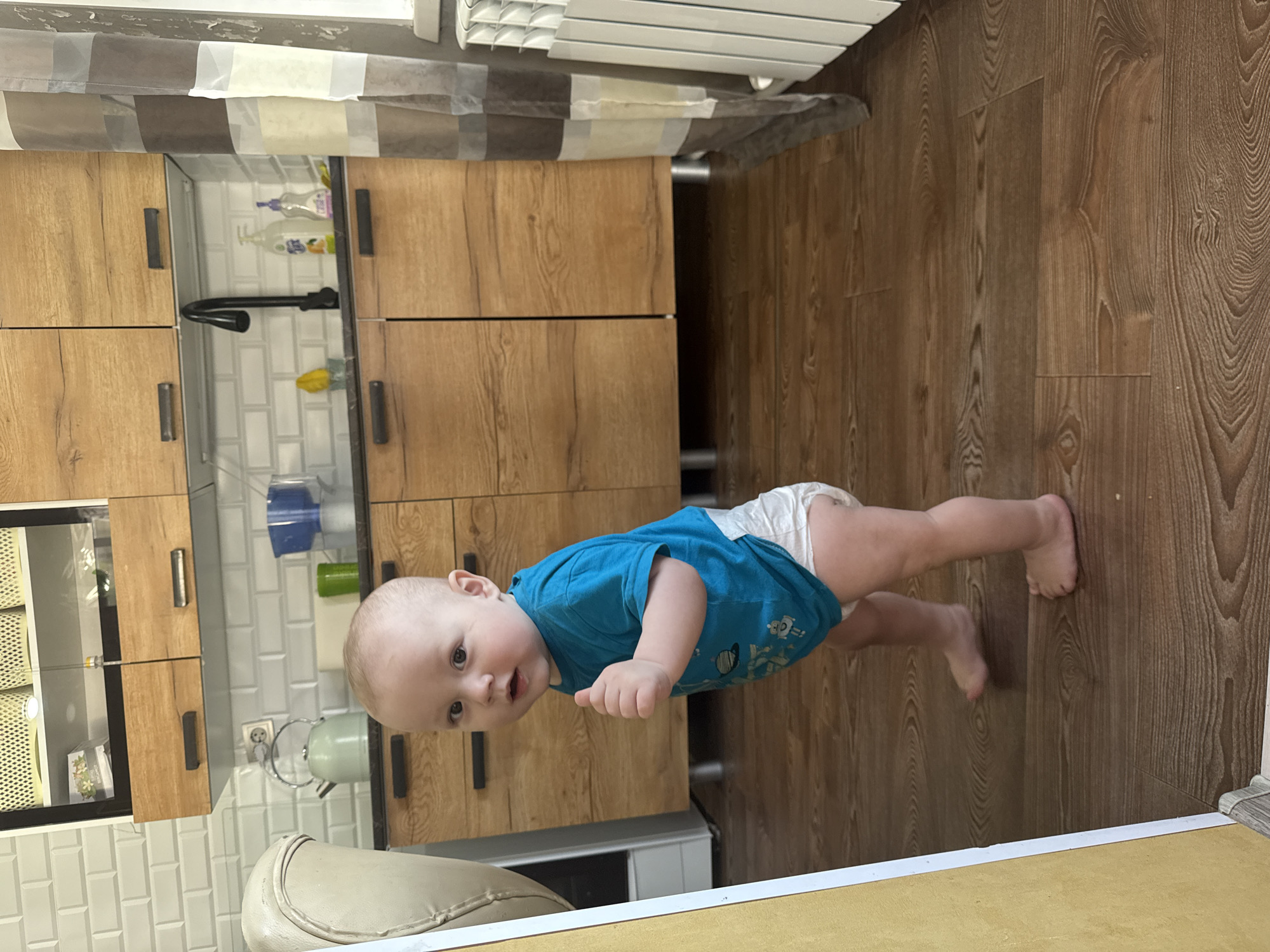 An infant is depicted standing barefoot on a brown wood-grain laminate floor in a kitchen located in the city of Кипчак, Russia. The infant, wearing a blue short-sleeved t-shirt with light-colored graphics and a white disposable diaper, faces the viewer with a slightly open mouth and wide eyes. Its left arm is bent with the hand partially open. The background consists of a modern kitchen with light-colored wooden upper and lower cabinets featuring dark horizontal handles, set against a white subway tile backsplash. A dark countertop holds a black gooseneck faucet, two soap dispensers, a yellow sponge, a light green kettle, a blue plastic container, and a green cup. Upper cabinets with glass fronts display woven baskets and a package. To the immediate left foreground, an off-white chair and a vertical yellow panel are partially visible. To the right, a multi-striped curtain in shades of brown, beige, and white hangs, partially obscuring a white radiator by a window.