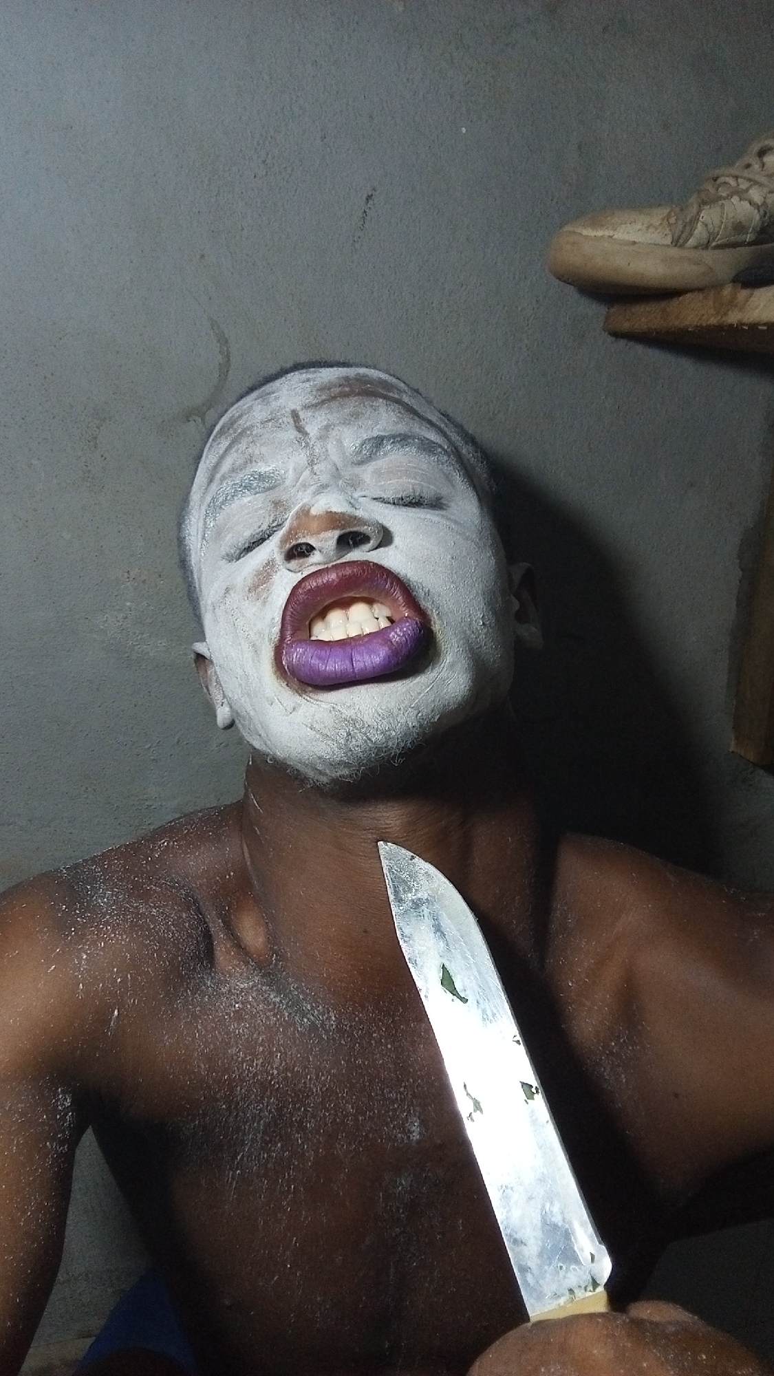 The image captures a close-up of a person with dark skin, their face dramatically covered in a white, powdery substance. Their eyes are squeezed shut, and their mouth is wide open, revealing white teeth and lips painted a vivid purple or dark red. The expression appears intense and theatrical.
The individual is bare-chested, with traces of the white substance visible on their shoulders and upper chest. They are holding a metallic knife with a light-colored handle, positioning the blade close to their throat or collarbone area, pointing upwards. Small green specks are visible on the blade of the knife, suggesting residue or a natural element.
The setting appears to be indoors, against a plain, dark gray wall with some minor imperfections. In the upper right corner of the frame, a pair of worn, light-colored shoes are resting on a wooden shelf or ledge. The lighting is dim and casts shadows, highlighting the contours of the person's face and body. The overall impression is one of a staged or performance art piece, evoking strong emotions, possibly of pain, ecstasy, or defiance. There is no visible text in the image.