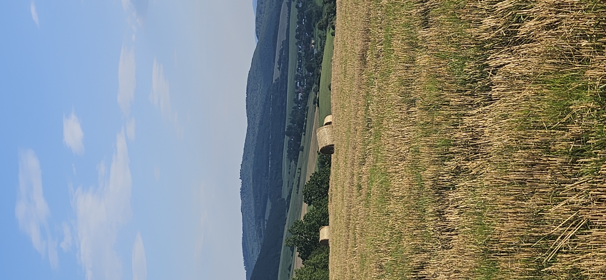 The image shows a rural landscape in Beňadikovce, Slovakia. The foreground is a field of dry, cut stalks of grain, with some green grass growing between them. In the midground, there are several round bales of hay scattered across a green field. Behind the hay bales, there is a line of trees, and beyond that, a small village is nestled in a valley. The village features houses with red roofs and other structures. Further in the background, rolling hills covered in dark green trees rise up against a clear blue sky with a few wispy white clouds. The overall scene depicts a harvested agricultural landscape under a bright, sunny sky.
