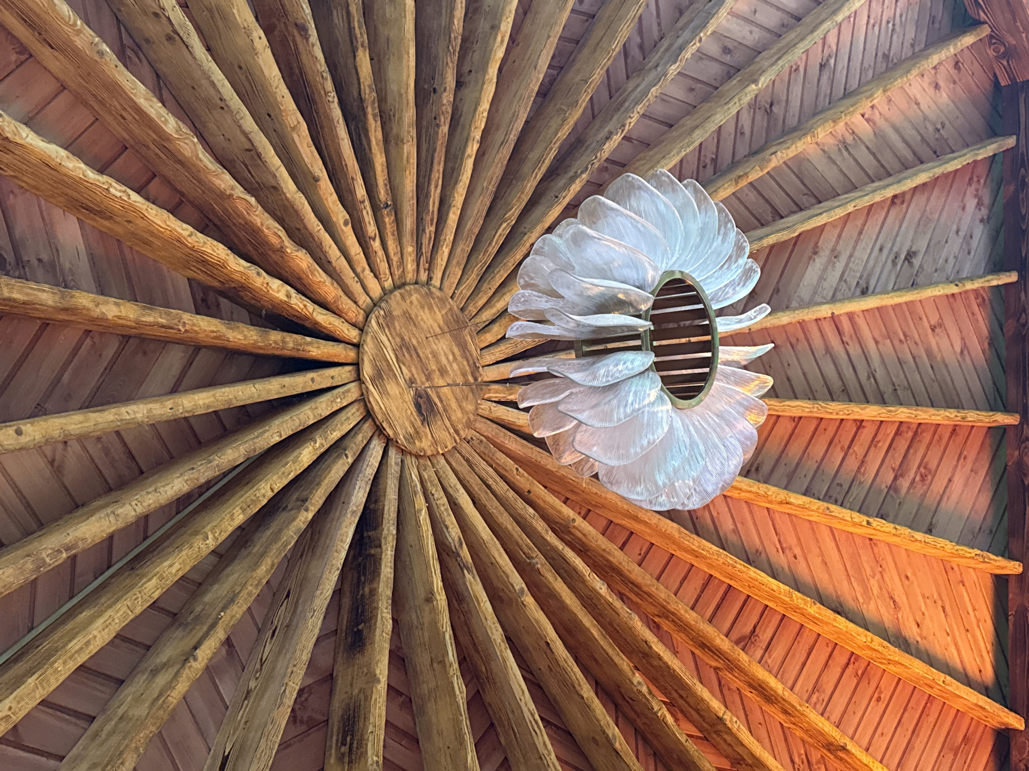 The image displays the interior of a wooden structure, likely a gazebo or pavilion, with a conical roof made of exposed wooden beams radiating from a central circular wooden element. The beams are rough-hewn and natural, with visible grain and imperfections, and they are supported by a wooden planked ceiling. A striking, modern chandelier with translucent, petal-like elements hangs from the center of the roof. The warm lighting creates an inviting atmosphere. The structure is located in Votkinsk, Russia. There are no people visible in the image.