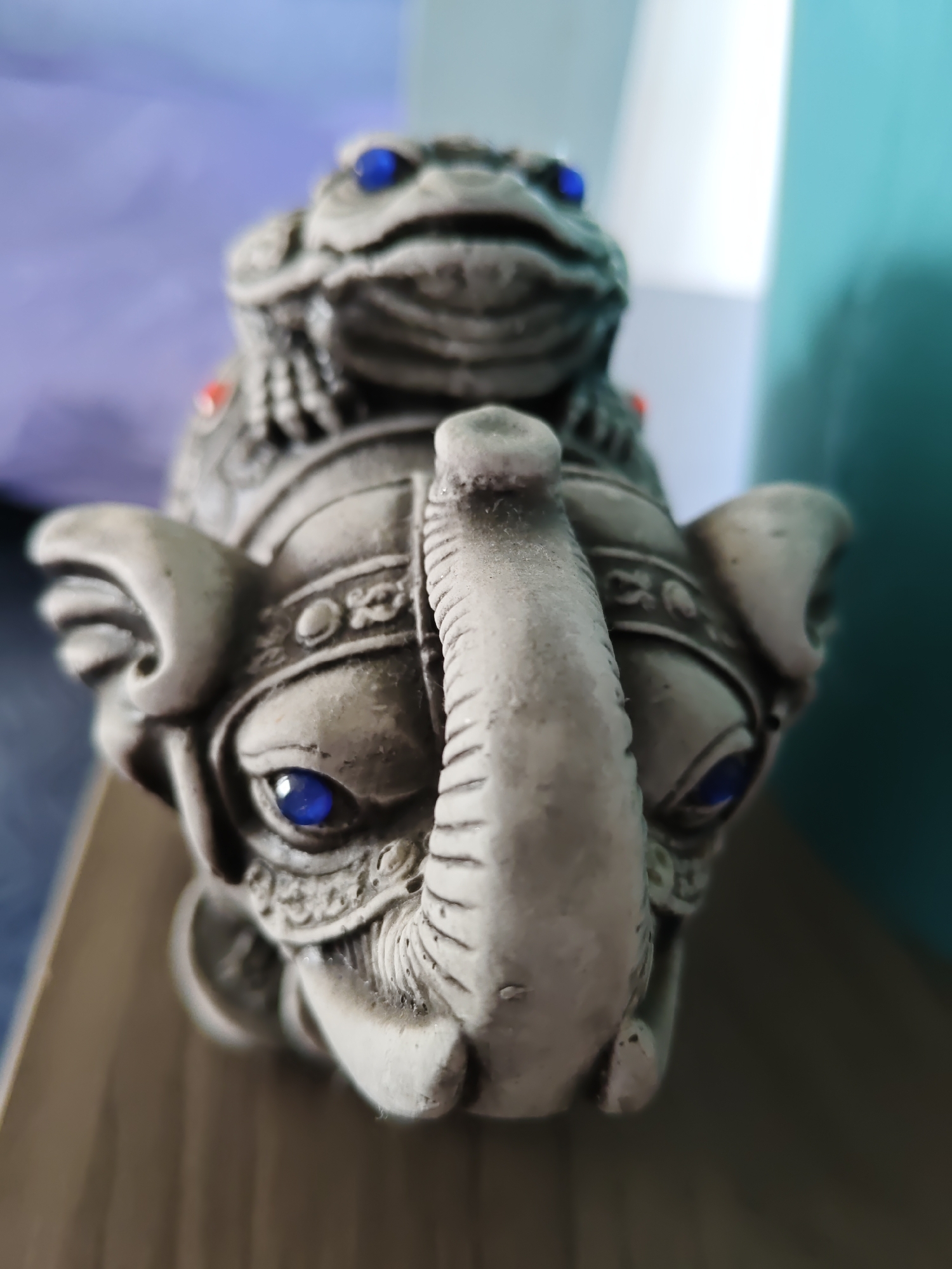 A close-up, high-angle shot focuses on a small, grey, stone figurine of an elephant with a frog on its head. The elephant has large, detailed ears, a textured trunk with prominent tusks, and intricate carvings on its forehead, including what appears to be a decorative headdress. The eyes of both the elephant and the frog are made of bright blue beads, adding a stark contrast to the muted grey of the figurines. Small red accents are visible on the elephant's headdress. The figurine rests on a surface that appears to be made of wood, with a visible grain. The background is blurred, showing a transition from a light purple to a teal color. There is no indication of the city of Rostov-on-Don, Russia, or any other specific location.