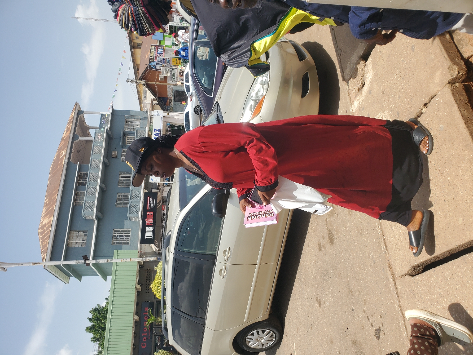 The image shows a street scene in Jos, Nigeria. A woman in a red dress, black leggings, and a black baseball cap is walking on the sidewalk. She holds a white bag and a pink notebook labeled "10inOne Notebook". A tan minivan is parked on the street next to her. In the background, there are buildings, shops, and a variety of goods hanging for sale. Several cars are parked on the street. Other pedestrians are visible in the scene. The weather appears to be sunny.