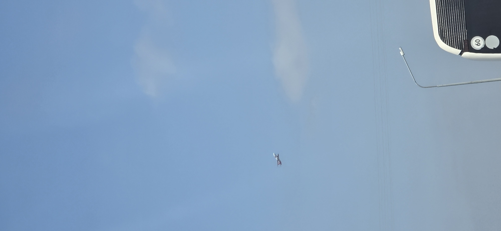 The image is a vertical, full-frame shot taken from a low angle, looking up at a clear blue sky. In the mid-left section of the sky, a small, red and white airplane is visible in flight. The plane appears to be a propeller-driven aircraft with its nose pointed slightly upward. Along the bottom of the frame, a portion of a white bus is visible on the right side, with a street lamp arching over it. The bus has a circular sign with the number "60" inside, indicating a speed limit. Faint horizontal lines, likely power lines, are visible across the sky. The scene is consistent with a view from a road or roadside area in Mineralnye Vody, Russia, likely during daylight hours given the bright sky.