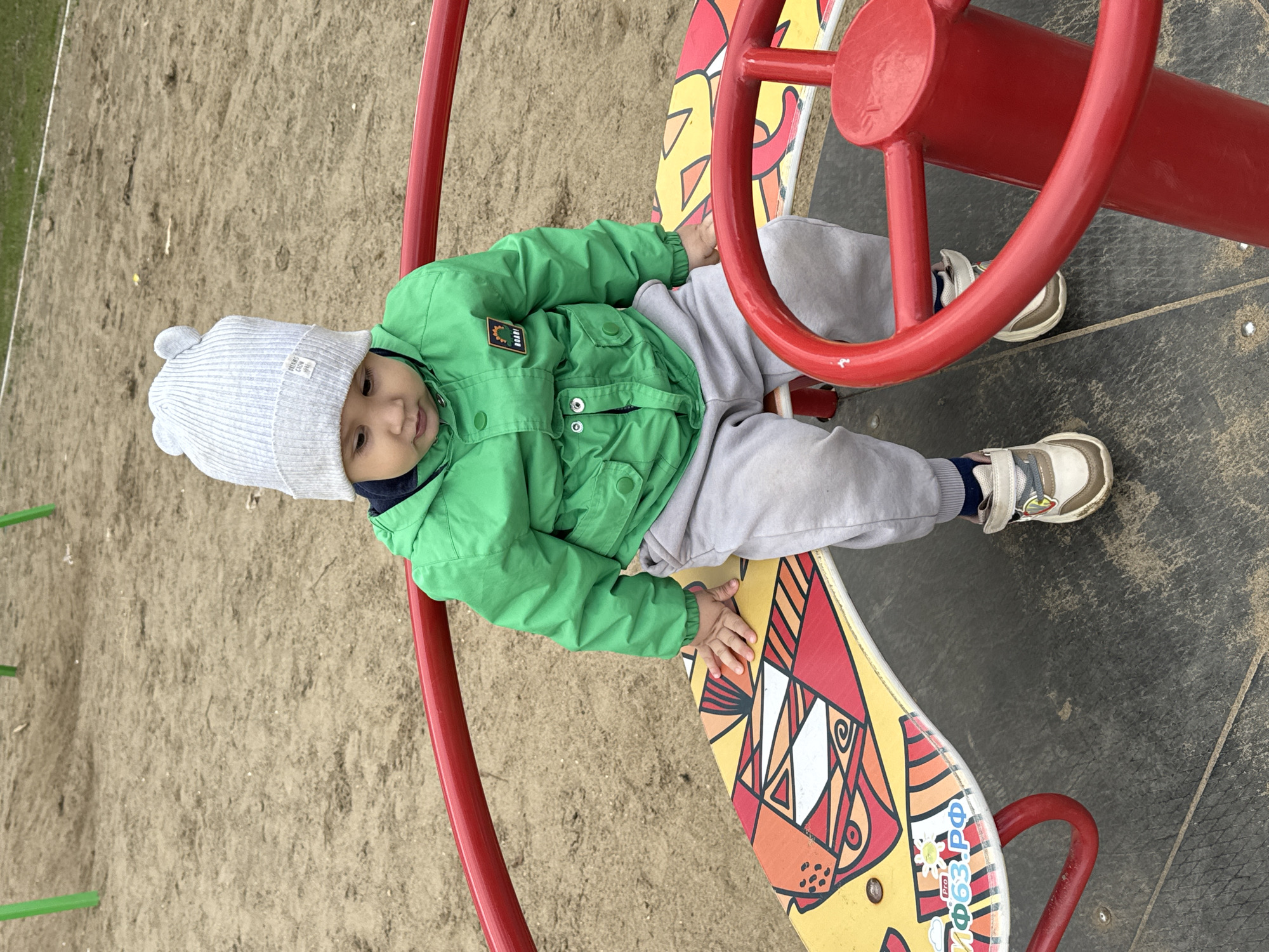 A young child is seated on a colorful playground spinner in Chaikovsky, Russia. The child, likely a toddler, wears a light grey ribbed beanie with two small pom-poms and a white label, partially displaying "HELLO CREW 1991". They are dressed in a bright green zip-up jacket, light grey sweatpants, and light-colored sneakers. The child looks directly at the viewer with a calm, slightly smiling expression.
The playground equipment features a vibrant yellow base with red, orange, and white geometric patterns, reminiscent of a stylized animal or fish. Integrated into the spinner are red metal handlebars and a prominent, round red steering wheel-like handle. Visible on the yellow base are the Cyrillic characters "ПРО" (PRO) and "63.РФ" (.RF), along with a small sun icon. The child's hands rest gently on the patterned base.
The outdoor setting includes light brown sandy ground on one side and a dark grey, textured rubber safety surface on the other, typical of a playground environment. In the background, green grass and a faint white line, possibly from a sports field, can be seen. The soft, diffused lighting suggests an overcast day, and the child's warm clothing indicates cool weather.