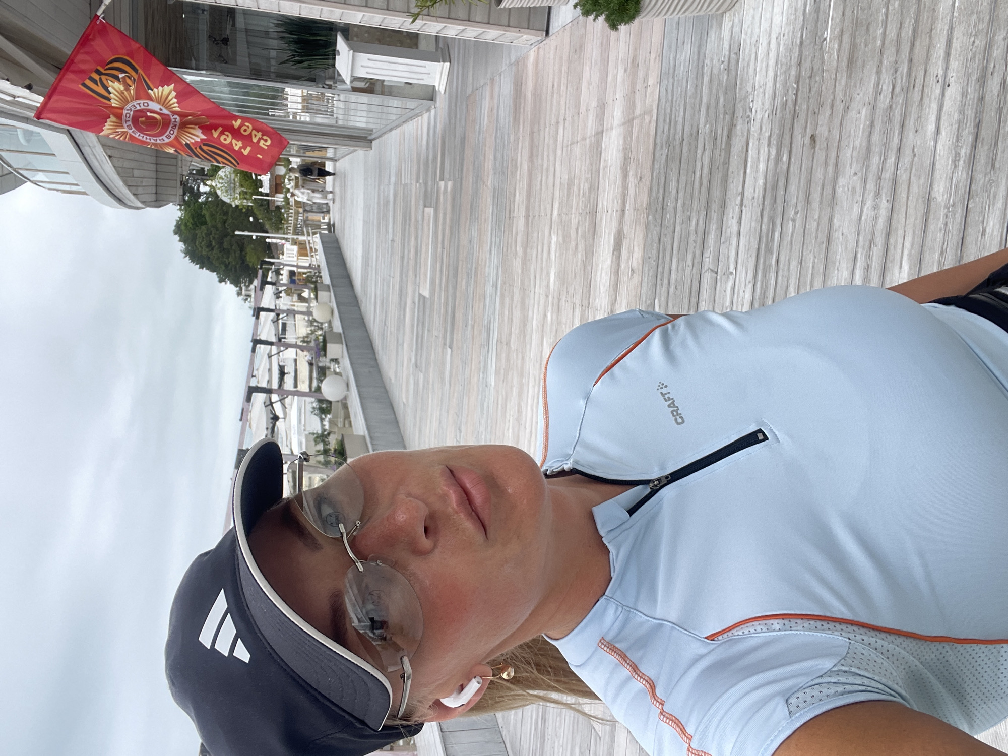 A selfie shows a woman wearing a dark-colored baseball cap with an Adidas logo, a light blue Craft athletic shirt with an orange trim, and rimless glasses. She has short blonde hair and a white earbud in her ear. Behind her is a wooden boardwalk with metal railings and decorative lights, and a building with white walls. A red flag with a Soviet military emblem hangs from the building. The location is Sochi, Russia.