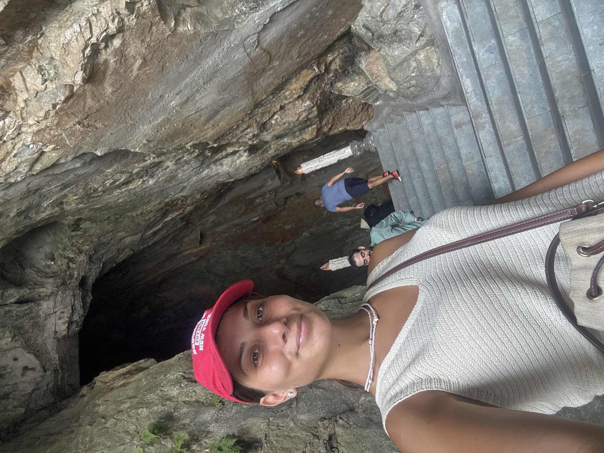 A woman wearing a red baseball cap, a white top, and a pearl necklace stands facing the camera, apparently taking a selfie at a stone cave entrance with grey stone steps leading up to it. A dark cavern is to her left. In the background, two men are visible: one in a blue shirt and dark shorts is walking down the stairs, and the other is wearing a green shirt and standing closer to the woman. Two white, cylindrical architectural features are positioned further back in the cave near the man in the blue shirt. The image appears to have been taken from an unusual angle, and the location is likely the Marble Mountains in Đà Nẵng, Vietnam.