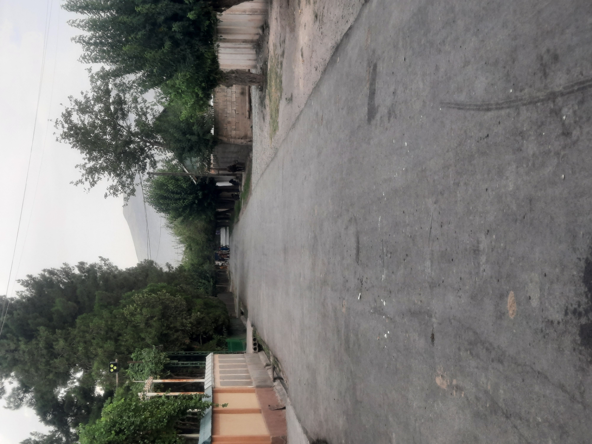 The image depicts a street scene in Восеабад, Tajikistan. The road is paved and appears to be in moderate condition, with some debris scattered on the surface. On the left side of the road, there is a low, tan-colored wall with a decorative pattern and a fence. Adjacent to the wall, a lush green tree with extensive foliage stands tall. Further down the street, several people, including children, are visible walking with bicycles. On the right side of the road, a brick wall runs along the perimeter of properties, with trees planted sporadically. The background shows a hazy mountainous landscape under an overcast sky. A traffic light with three green lights is visible above the wall on the left. The overall atmosphere suggests a residential area in a rural or semi-urban setting.
