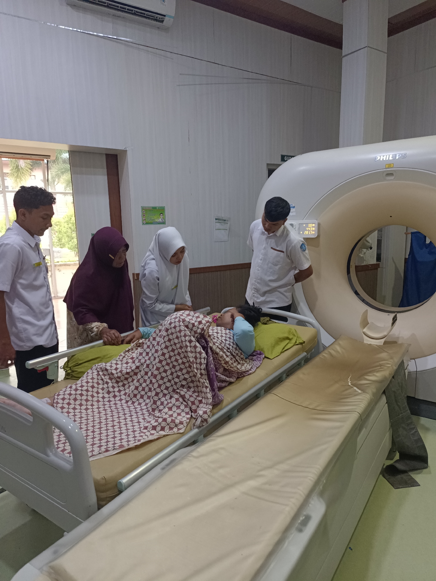 In a hospital room in Mesjid Reuntoh, Indonesia, a patient lies on a gurney, covered with a patterned blanket. The patient is positioned next to a large, white MRI machine, with the circular opening of the machine visible. Three individuals are present in the room, seemingly attending to the patient. Two are medical professionals, one a male in a white uniform and the other a female in a white hijab and uniform, observing the patient. A third individual, an older female wearing a dark hijab, stands beside the gurney, appearing to be a family member or caregiver. The room is lit with overhead fluorescent lighting and features an air conditioning unit mounted on the wall. Medical equipment, including another gurney, is also visible in the background.