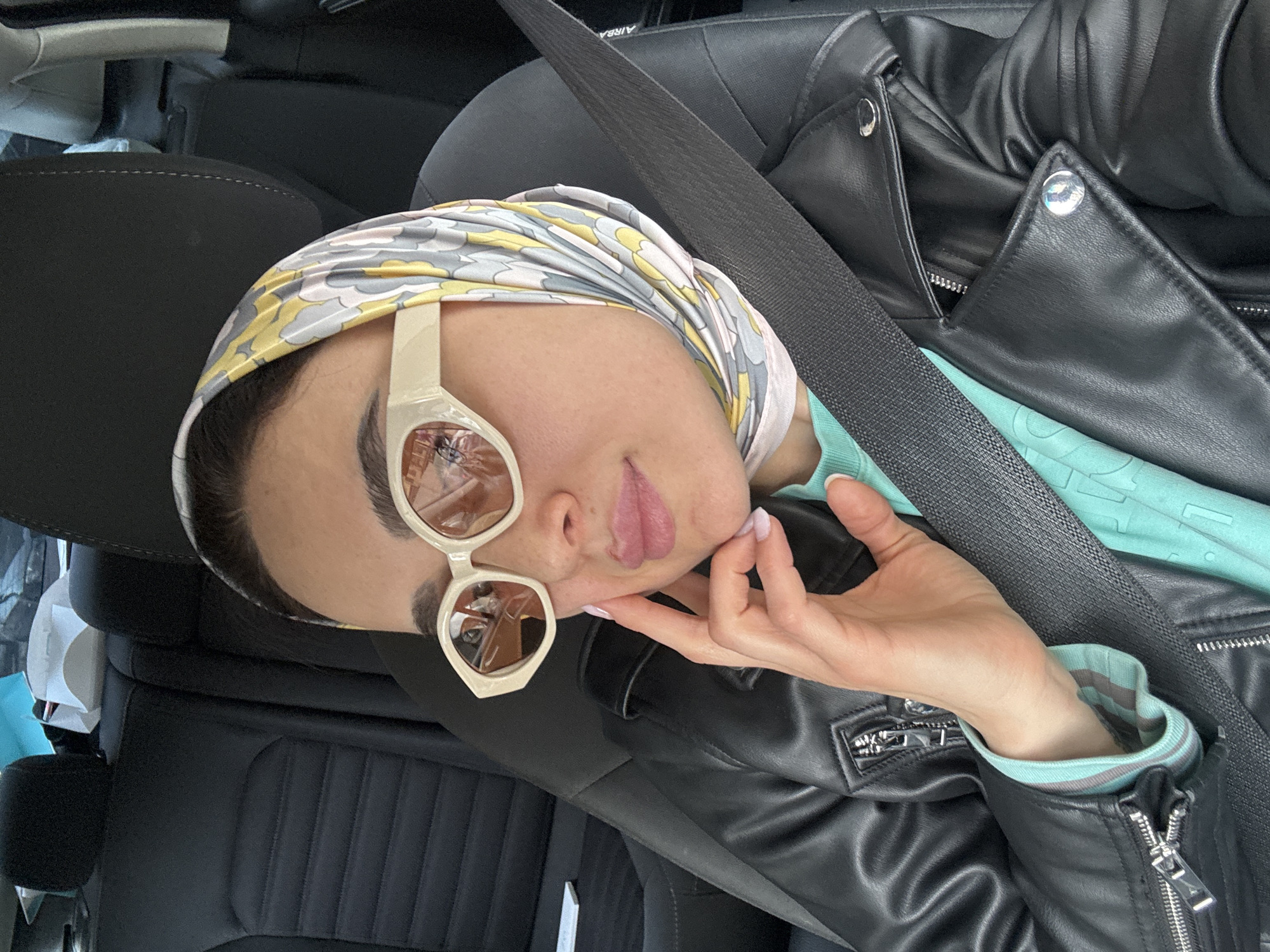 A person wearing sunglasses, a patterned headscarf, a light blue top, and a black leather jacket is in a car. They have their hand near their face and appear to be posing. The person is wearing a seatbelt. The setting suggests the inside of a vehicle, potentially in transit. It is not possible to determine the location in the city of Tbilisi, Georgia based on the image.
