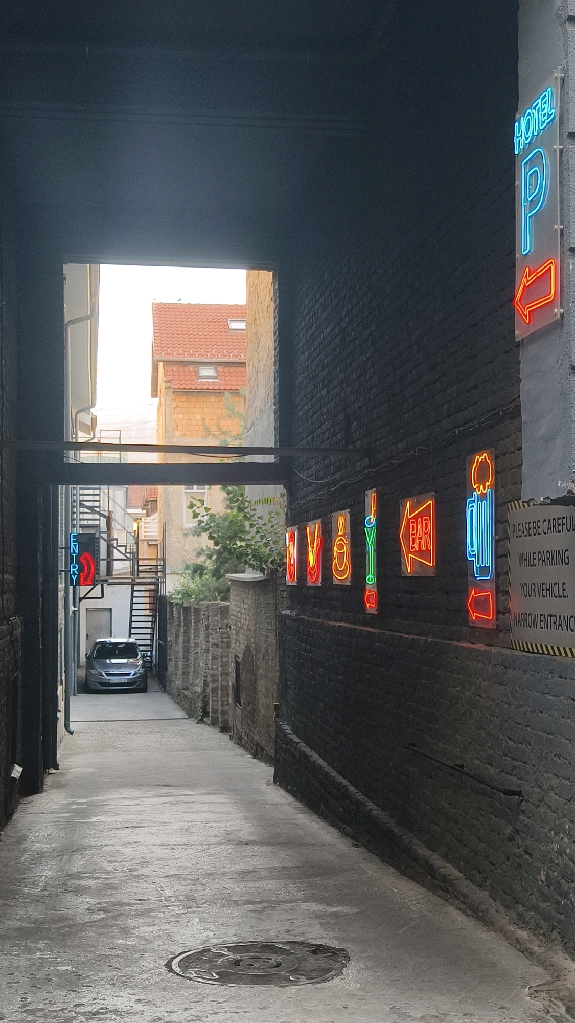 The image depicts a narrow alleyway in Belgrade, Serbia, with a car parked at the far end. The alley is lined with brick walls, and several neon signs are visible. On the right wall, a prominent blue and red neon sign reads "HOTEL P" with an arrow pointing left, indicating parking. Further down the right wall, a series of colorful neon signs depict various icons, including a coffee cup, a martini glass, and what appears to be a beer mug. There is also a neon sign for a "BAR" with an arrow. On the left wall, a red neon sign reads "ENTRY" with a red arrow pointing downwards. The alley floor is paved and has a visible manhole cover. Buildings are visible in the background, including one with a red tiled roof. The overall impression is one of a vibrant, perhaps nightlife-oriented, area within the city.