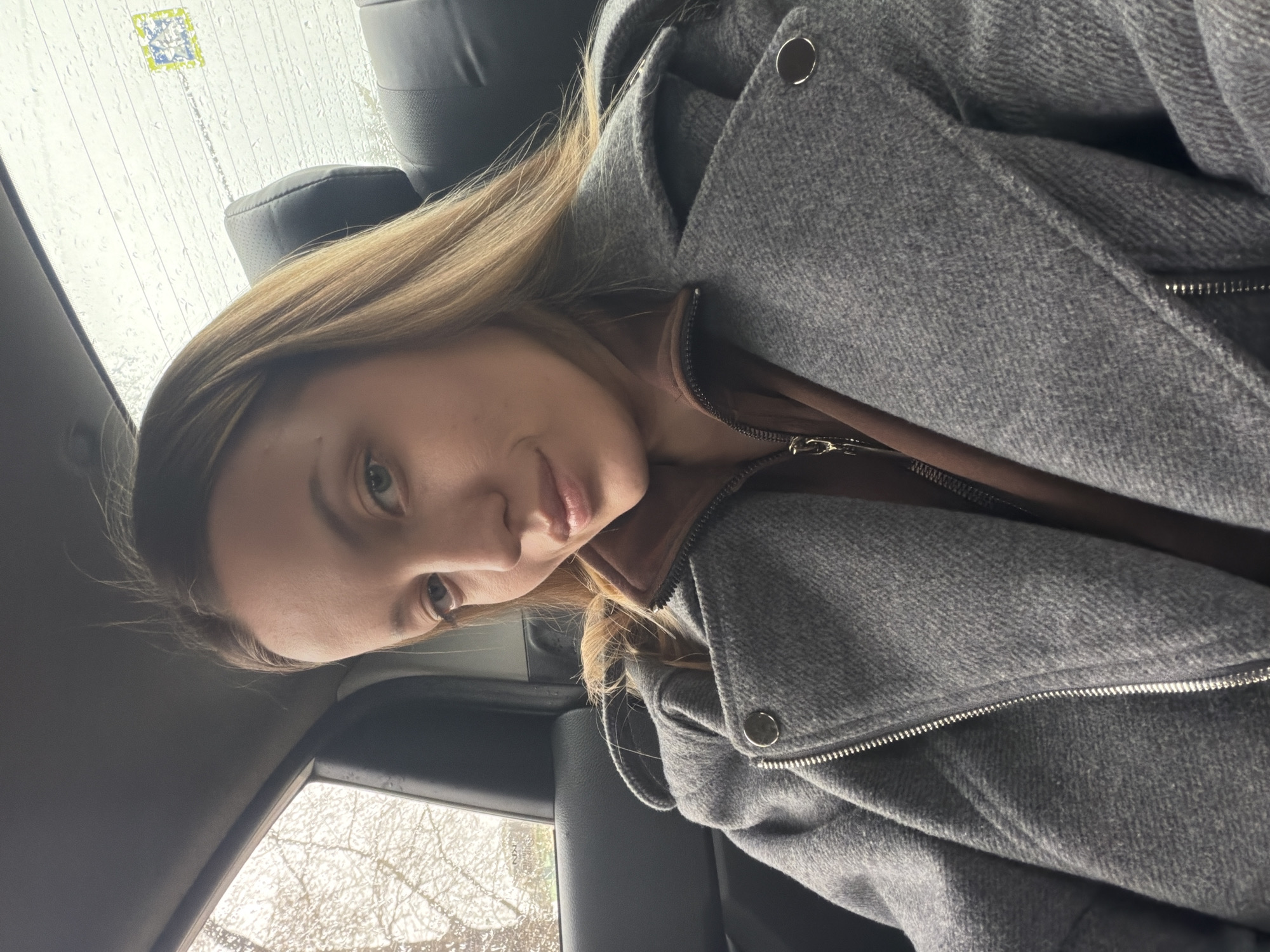 The image is a selfie taken inside a vehicle. A woman with blonde hair and fair skin is looking at the camera. She is wearing a gray hooded jacket over a brown top. The vehicle appears to be stationary, as seen by the raindrops on the window to the left. Outside the window, bare trees are visible, suggesting a cold season. A sticker is seen at the corner of the windshield. There is no specific indication that this image was taken in Klin, Russia.