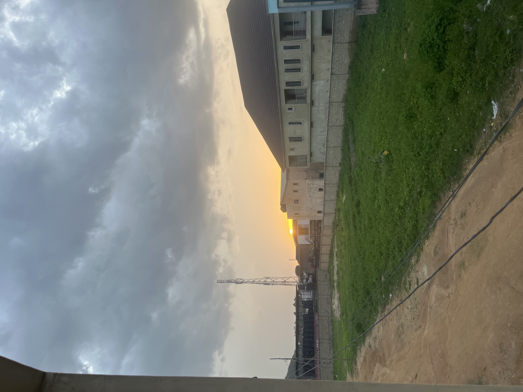 The image depicts a sunset over Gura Top, Nigeria. The sky is filled with dramatic, heavy clouds, with the sun peeking through the horizon, casting a warm glow on the scene. In the foreground, a dirt path runs alongside a grassy, undeveloped lot bordered by a concrete block wall. Beyond the wall, two multi-story buildings with tiled roofs are visible. To the left, a tall telecommunications tower stands prominently against the twilight sky. Structures that appear to be large storage containers or sound equipment are stacked in front of a building to the left of the tower. The overall atmosphere is one of urban development in a somewhat unfinished state, set against a natural and atmospheric sky.