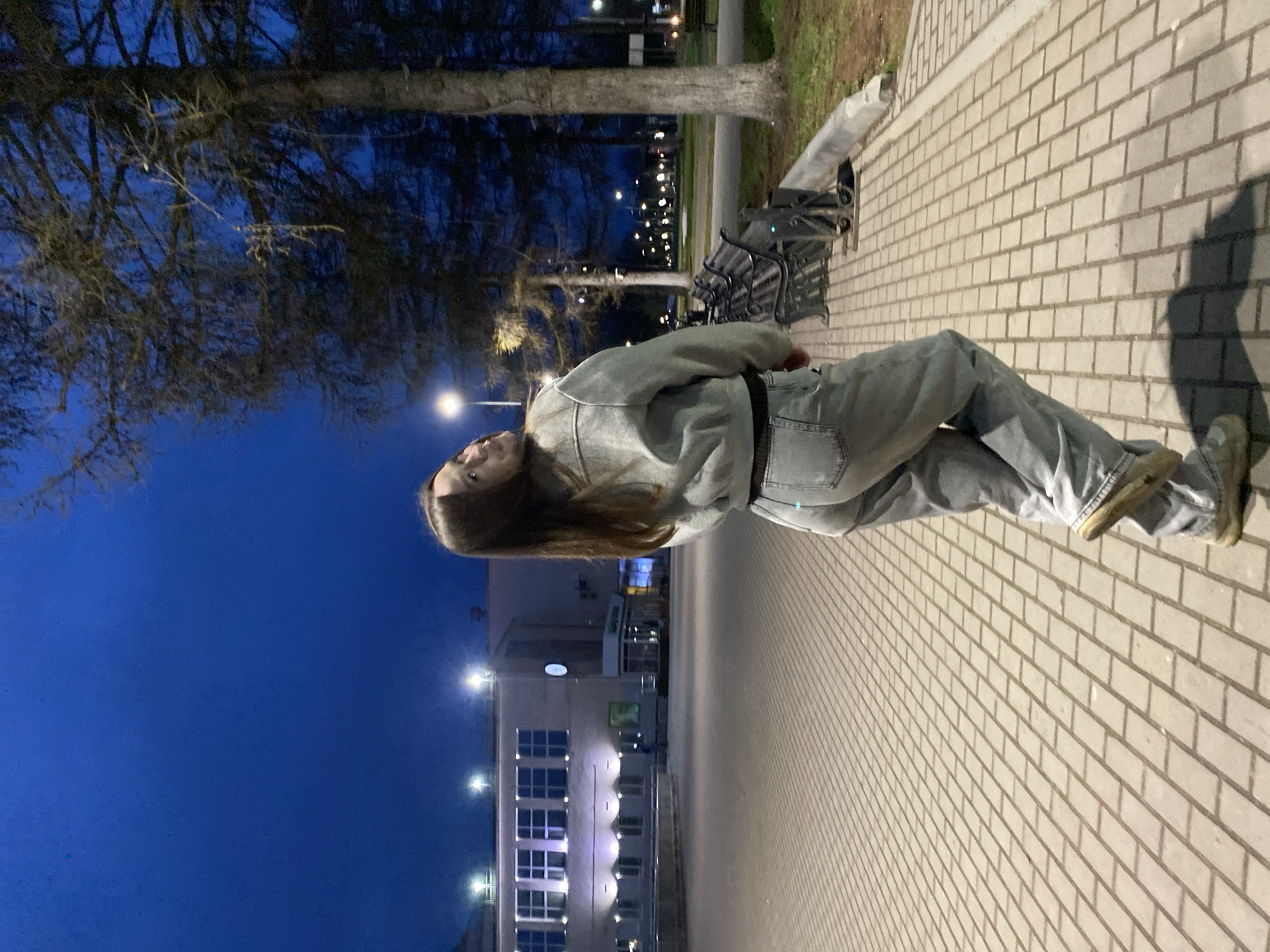 This image depicts a young woman standing on a paved pathway during the evening or night in Chaikovsky, Russia. She is positioned towards the center-right of the frame, looking slightly upwards and to her right with a neutral expression. She has long, straight, dark hair and is wearing a light grey or faded denim jacket, light-colored denim jeans, and light brown or beige shoes. A dark belt cinches her waist, and a dark mask or scarf is visible around her neck.
The setting appears to be an urban or park area. The pathway is made of light-colored rectangular paving stones, stretching into the background. To her left and behind her, several bare trees with dark, intricate branches stand out against a deep blue, dark sky, suggesting a cooler season like late autumn or winter. A prominent tree trunk is visible near a dark metal park bench with slatted seating. In the background, beyond the trees, a multi-story building with numerous illuminated windows can be seen. Multiple streetlights are also visible, casting artificial light across the scene. The overall atmosphere is quiet, and the scene is illuminated solely by artificial light sources. No visible text is present in the image.
