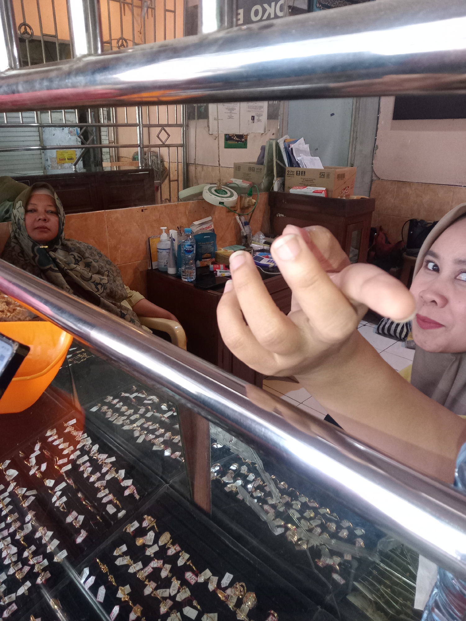 This is a medium shot, eye-level interior photograph taken in a shop in Borobudur, Indonesia, likely a jewelry store. In the foreground, a reflective metal bar runs horizontally across the frame, obscuring a portion of the view. Below the bar, a glass display case is filled with numerous rings, each with a small white price tag attached. The rings appear to be made of gold and are arranged in black velvet trays.
To the left, partially visible is a woman wearing a patterned headscarf, looking directly at the camera. She is seated in a chair with an orange bowl to her left. In the background, shelves are stocked with various items, including boxes and bottles. A wooden counter runs behind the seated woman.
On the right side of the image, a person's hands are in the foreground, reaching towards the camera. A woman wearing a light brown hijab is visible in the background on the right, looking slightly away from the camera. The overall environment appears to be a retail space with a focus on displaying jewelry. The lighting suggests an indoor setting with possibly artificial light sources.