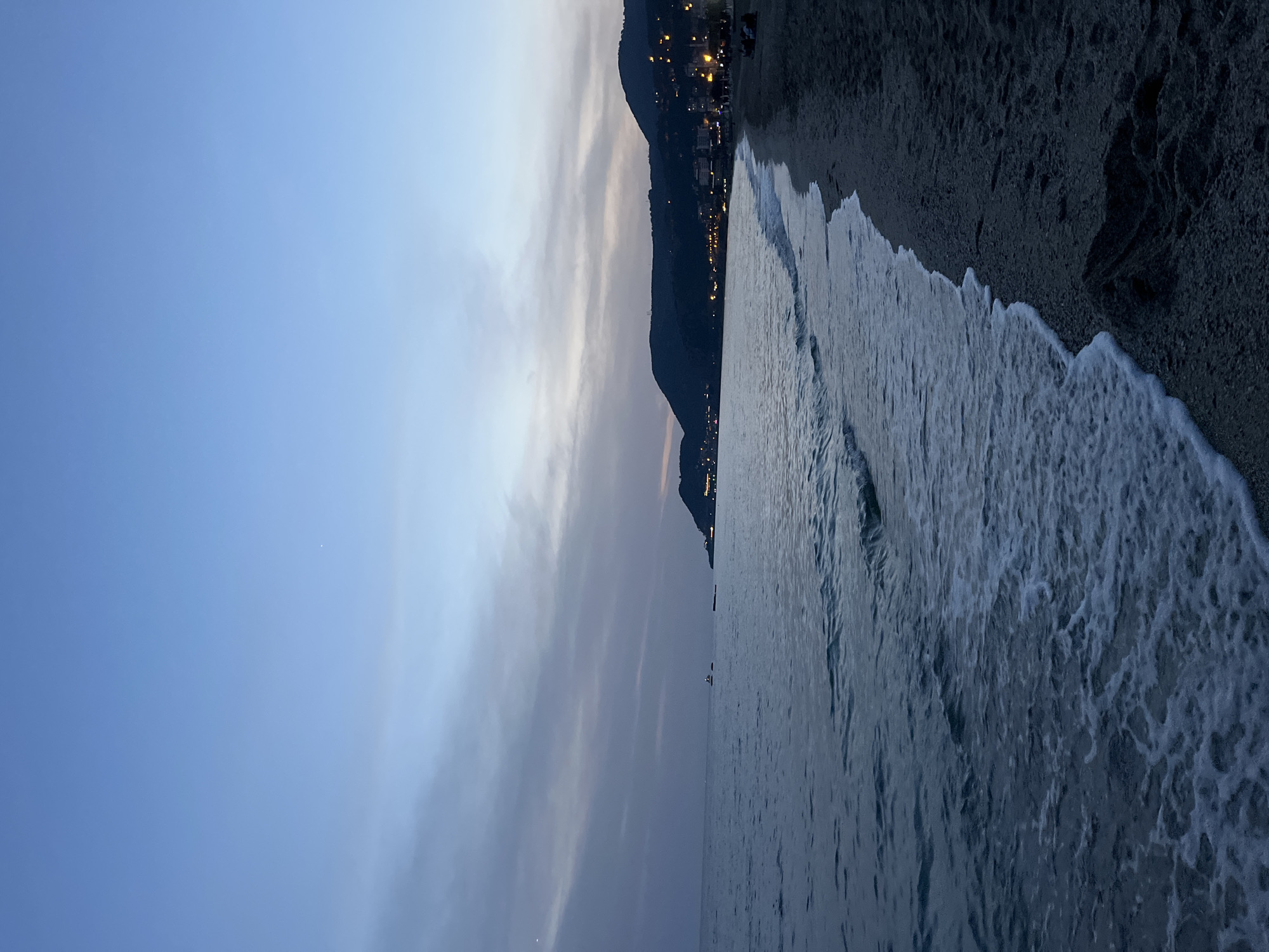 A nighttime shot of a beach and sea in Alanya, Turkey, showing a dark sandy beach on the upper right of the frame. Waves wash towards the beach. A mountain range with city lights runs down the center of the shot. The sky, visible on the left, is blue, but lightens toward the horizon behind the mountain. There are some ships or boats visible in the distance between the camera and the mountain.