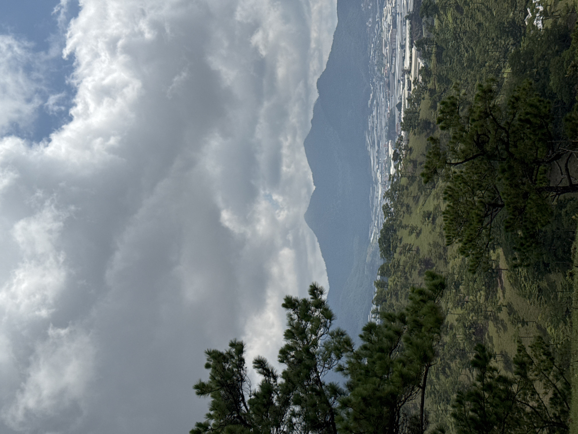 The image shows a mountain range in Lạc Dương, Vietnam. The left of the frame is dominated by a cloudy sky with patches of blue visible. To the right, a mountain rises. Its lower slopes are covered in dense green vegetation, including pine trees in the foreground. Beyond the mountain and extending into the distance, are what appear to be numerous white structures, possibly greenhouses or buildings. The landscape has a hazy, overcast atmosphere.