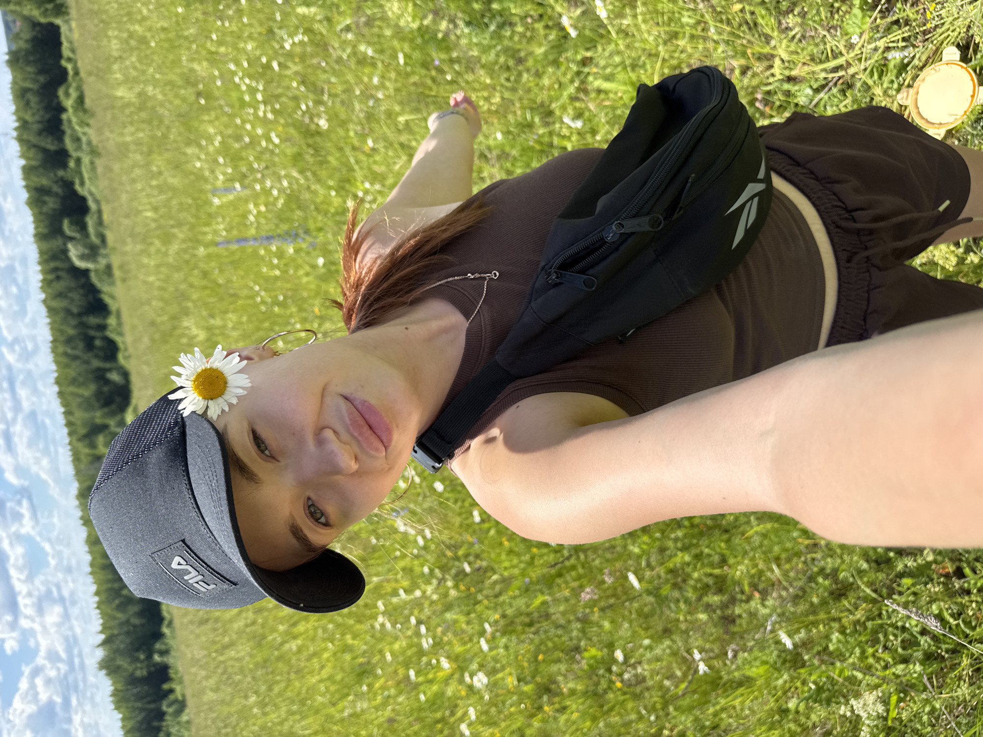 A young woman with red hair is taking a selfie in a grassy field. She is wearing a brown tank top, brown shorts, a black fanny pack, and a black baseball cap with a "FILA" logo. A daisy is tucked into the brim of her cap. She is smiling and looking at the camera. The field is filled with small white and yellow wildflowers, and a line of trees can be seen in the background, along with a blue sky with white clouds. The sun is shining, suggesting it is daytime. The overall mood is cheerful and relaxed, consistent with a warm, sunny day outdoors in a natural setting.