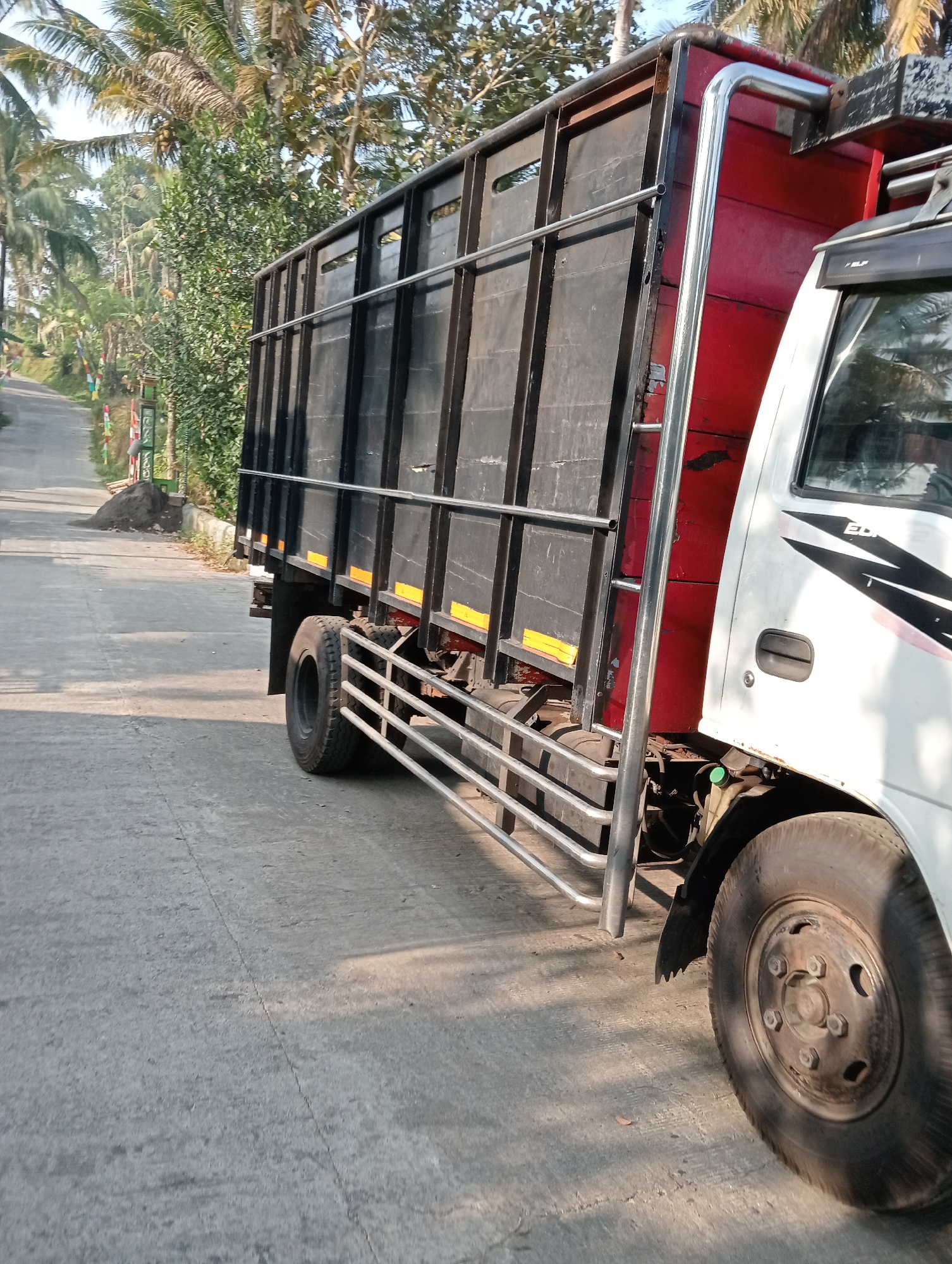 A side view of a white truck with a red cargo box is parked on an asphalt road. The truck is facing to the right of the frame, and its front is partially visible. The cargo box is black and appears to be made of metal. There are palm trees and other greenery on the left side of the road, which is lined with a small wall. The sky is clear and blue. The location is Krajan Kidul, Indonesia.