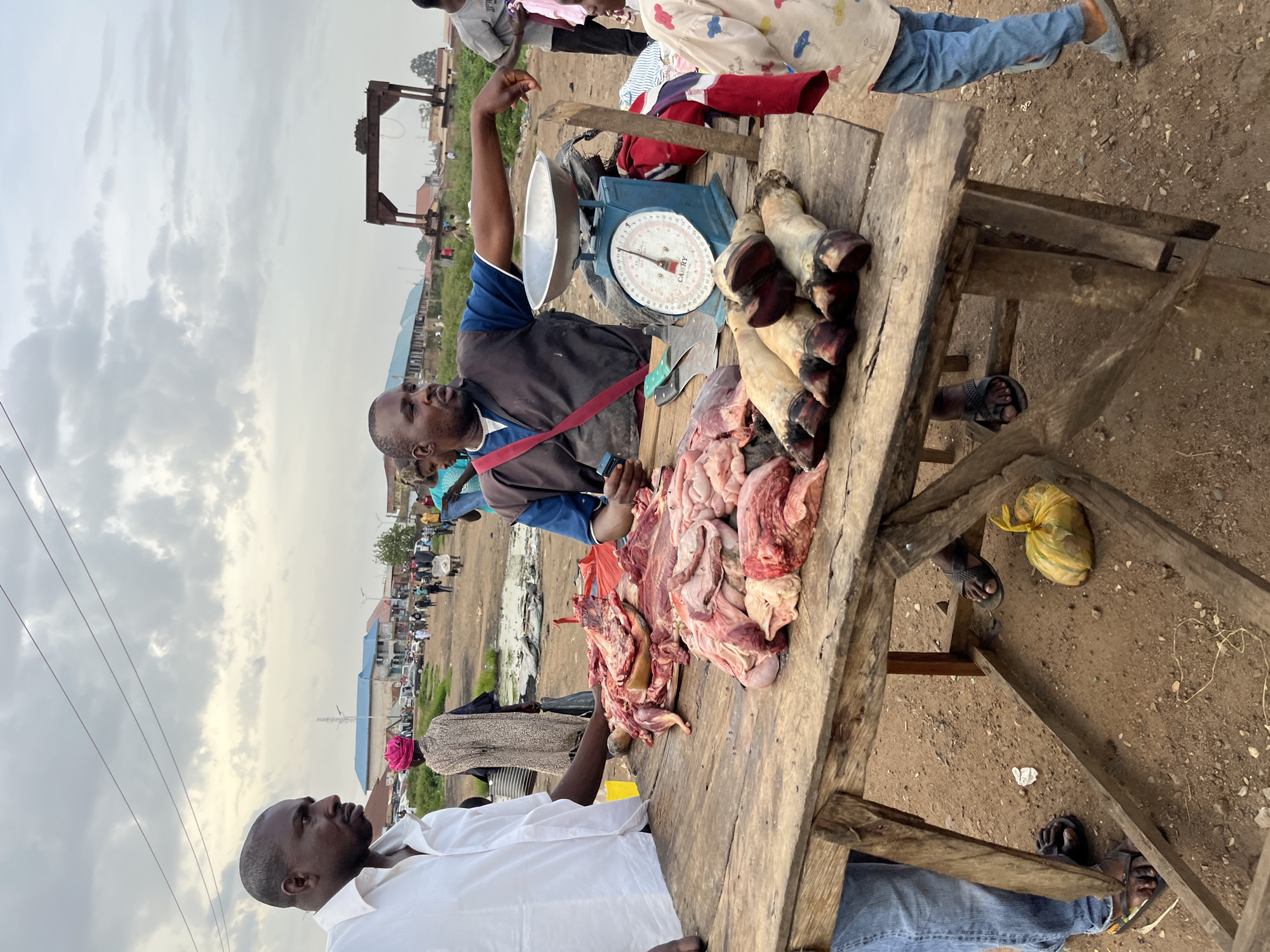 In Bukuru, Nigeria, a butcher stands behind a wooden table laden with raw meat, including various cuts and what appear to be animal trotters. He is gesturing with his right hand towards the meat, seemingly explaining something to a young person standing to his right. A blue scale is positioned on the table, ready for weighing. To the left of the table, a man in a white shirt is visible, his arm resting on the table. The setting is an outdoor market, with a dusty ground, a cloudy sky, and other market-goers and structures in the background, suggesting a bustling commercial area.