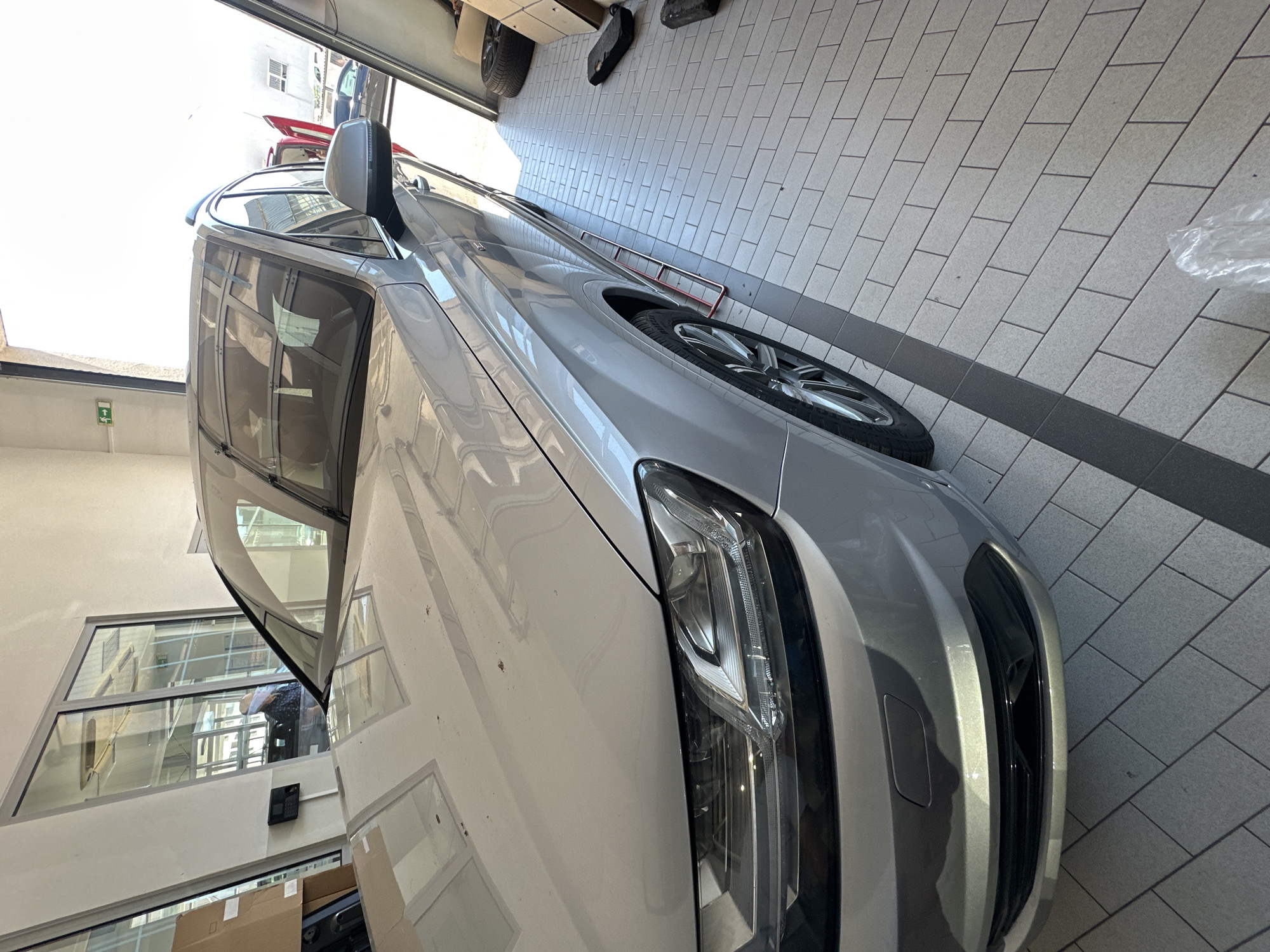 An outdoor scene shows the front left side of a silver Audi Q7 parked in a garage with the door open. The car has a shiny finish and is clean except for a few specs of dirt on the hood. The garage floor is made of light grey tiles with dark grey grout. There is a person in the background, looking out a window. The scene appears to be taken during the day, with bright sunlight streaming in from the garage door. There are some boxes and other items in the background, as well as some tools. The overall scene is well-lit and the car is the main focus.
