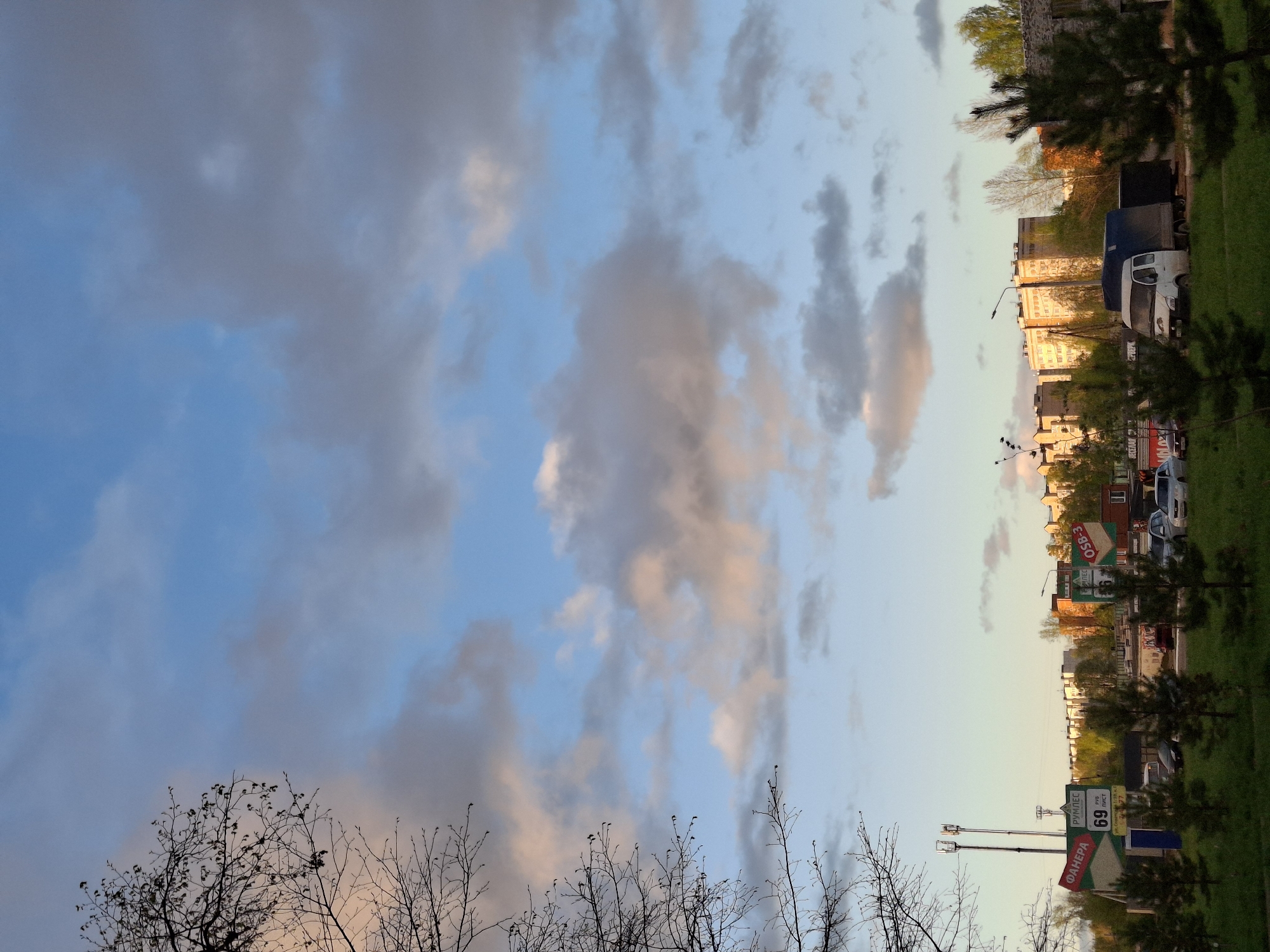 The image shows a cityscape, likely in the city of Chaikovsky, Russia, judging by the Cyrillic script on the signs. The view is directed upwards towards the sky, with a mix of blue sky and gray clouds dominating the frame. In the lower portion of the image, bare tree branches are visible against the sky. Along the right side, there's a line of buildings and trees, with glimpses of vehicles parked along the street. Signage with Cyrillic text indicates the presence of businesses or landmarks. Street lights and grassy areas are also visible along the roadside. The perspective is somewhat distorted, as if the camera was tilted upwards from a low angle.