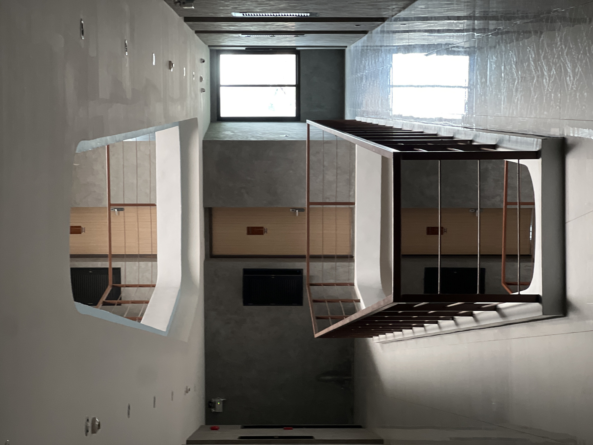 This is an interior shot of a multi-level modern building. The walls are predominantly gray, with smooth surfaces, possibly concrete or plaster. There's a large rectangular window near the top of the frame. A rectangular opening in the wall on the left offers a view into another area, reflecting the interior design.
A structure resembling a staircase or scaffolding is positioned diagonally in the center of the frame, possibly indicating ongoing construction. It is made of wood and metal.
The environment is clean and appears newly built or renovated. There are no visible people. The location is an urban interior, but specific details to identify the city are absent. The time of day is either daylight or illuminated by artificial light mimicking daylight, given the brightness through the windows.