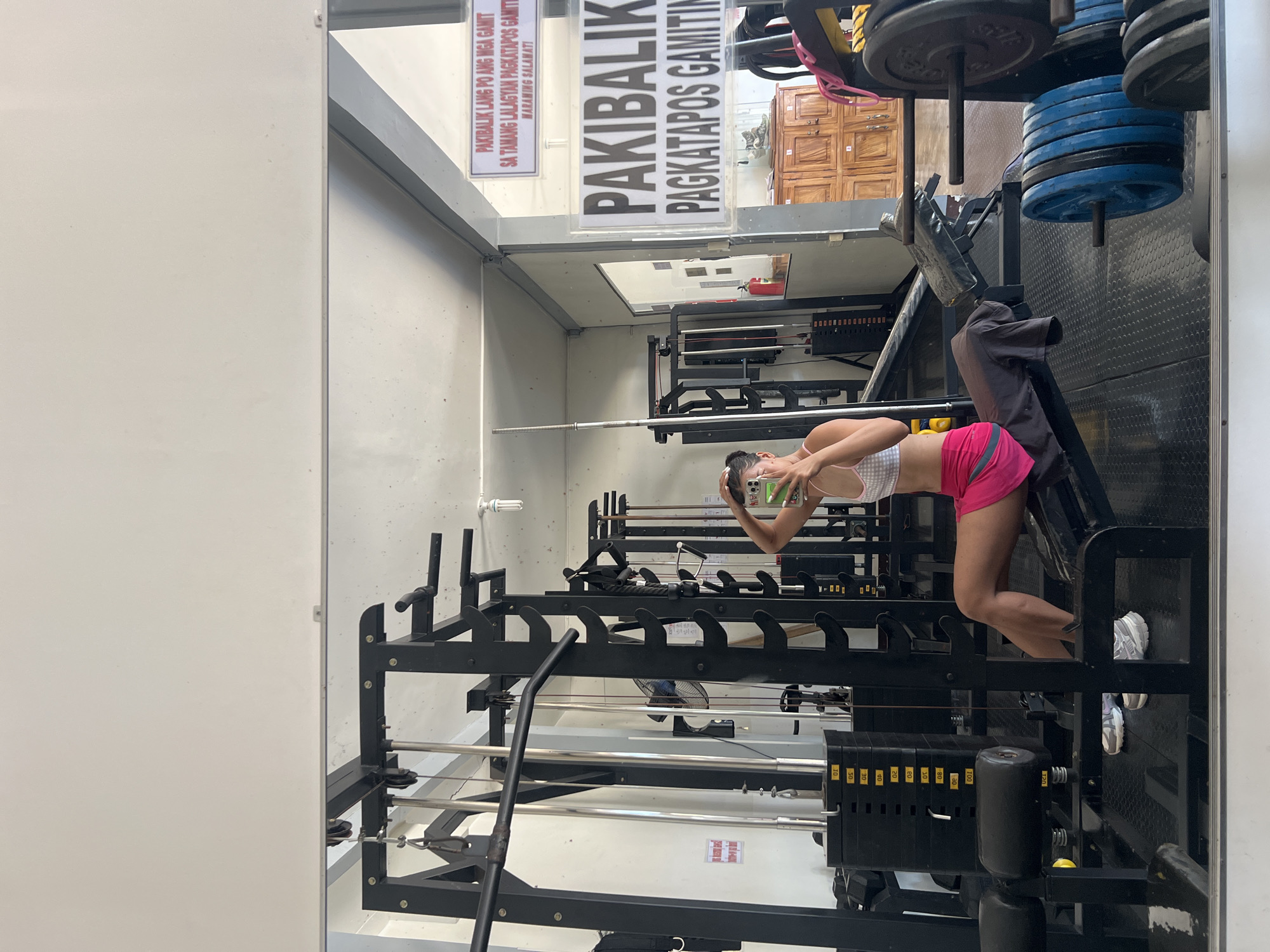 In a gym located in Cuyo, Philippines, a woman with dark hair pulled back is taking a selfie in a mirror. She is wearing a white and pink checkered sports bra and a bright pink skirt. She is seated on a weight bench, leaning back slightly, with her right hand touching her hair and her left hand holding a smartphone in front of her. The gym is equipped with various weight machines, including a leg press machine with stacked weights and blue weight plates, and a multi-gym apparatus with a weight stack. In the reflection, a sign with Filipino text that translates to "Please return equipment after use" is visible on the wall. The environment appears to be brightly lit with fluorescent lights.