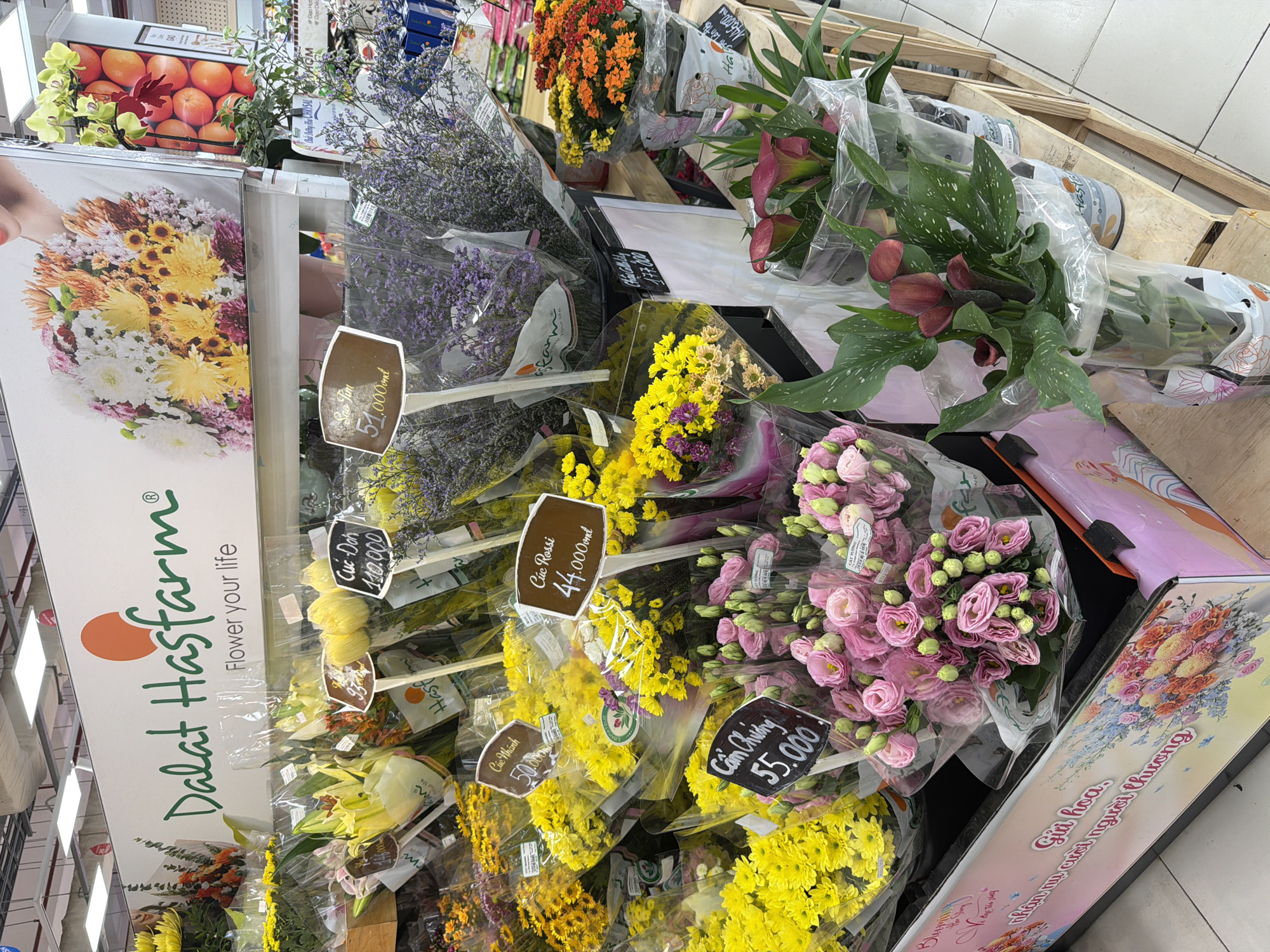 This image shows a display of fresh cut flowers for sale at a Đà Lạt Hasfarm store, likely in Đà Nẵng, Vietnam. The flowers are bunched and wrapped in clear plastic. Varieties include chrysanthemums, lilies, and calla lilies. Each bouquet has a small price tag written in Vietnamese. The sign displays the brand "Dalat Hasfarm" with the tagline "Flower your life". There is a poster with Vietnamese text that references sending flowers and seeing a loved one's smile.