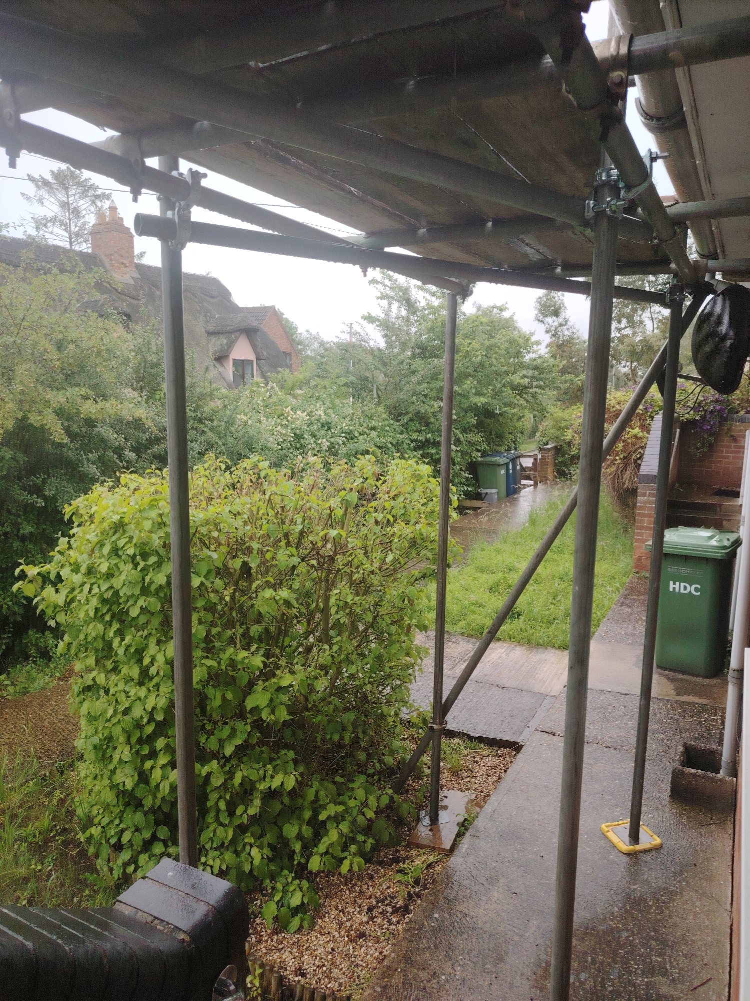 The image depicts an outdoor scene under scaffolding, likely in Huntingdonshire, United Kingdom, given the presence of an "HDC" branded green refuse bin. The scaffolding, constructed from metal poles and wooden planks, covers a walkway leading towards a building with a thatched roof. The weather appears overcast and wet, with visible water droplets on the scaffolding planks and a generally damp environment.
In the foreground, a dark, metallic object, possibly a piece of rubble or construction material, rests on the left. Adjacent to it is a dense, green bush. The walkway is made of concrete slabs, some of which are cracked or uneven. Further down the walkway, two green refuse bins are visible, one marked "HDC." Beyond the immediate area, lush green trees and vegetation create a dense, natural backdrop. In the distance, a traditional-looking building with a thatched roof and a red brick chimney is partially obscured by the foliage. The overall impression is of a residential or semi-rural setting undergoing some form of construction or maintenance work, set against a backdrop of natural greenery and traditional architecture.