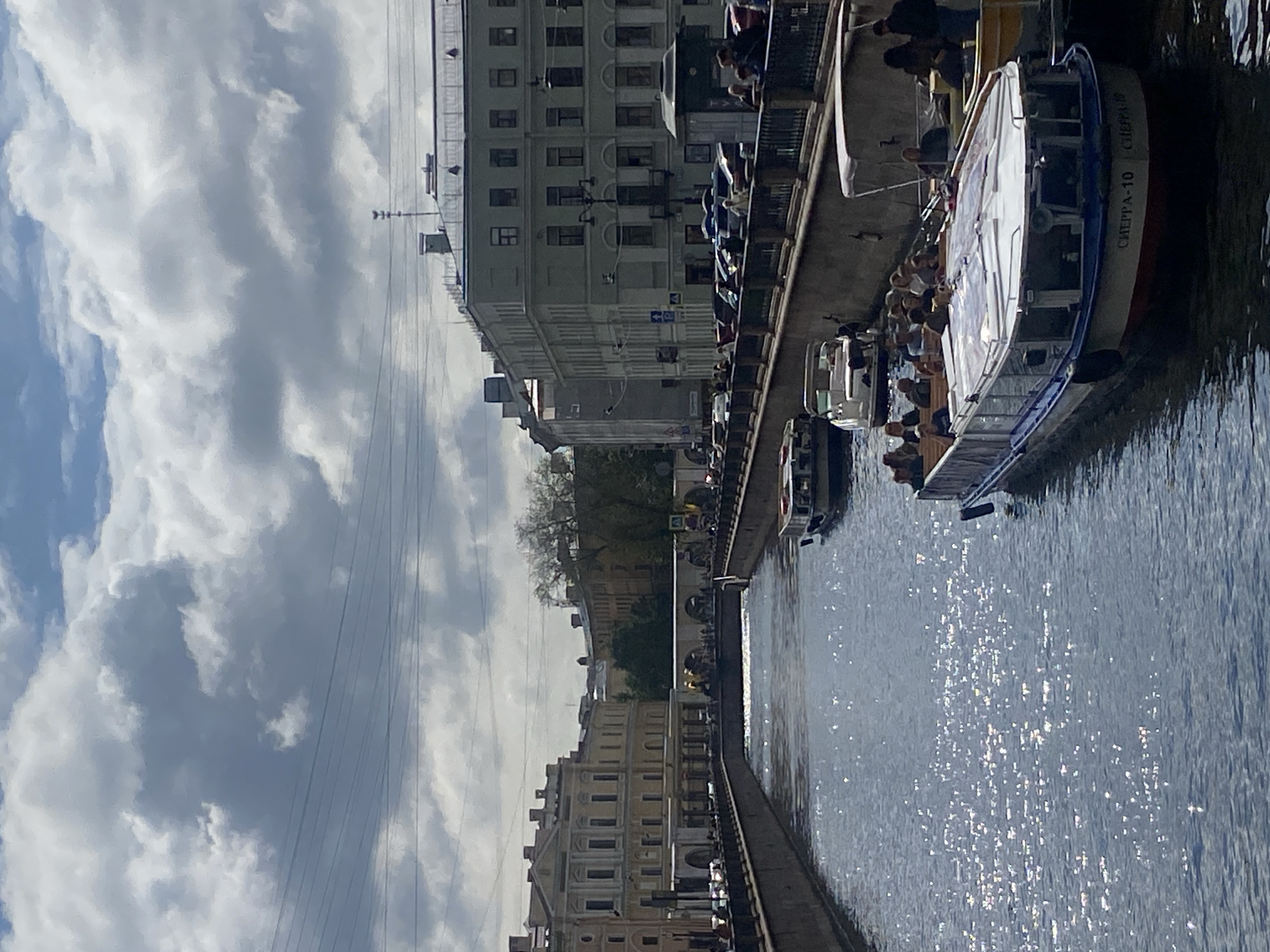 The image captures a bright, sunny day with scattered clouds in Saint Petersburg, Russia, likely showing a canal scene. Several boats are visible on the water. In the foreground, a large white passenger boat with a blue stripe along its hull is docked, with passengers seated inside and on the upper deck. Another smaller white boat with a canopy is moving away from the viewer on the left side of the canal. Further in the distance, another boat is also visible.
The canal is flanked by stone embankments, with a walkway or road running alongside. Buildings with distinctive architecture line the canal, characterized by their classical style with numerous windows, balconies, and decorative elements. Some buildings are light-colored, while others are a pale green. Electrical wires are strung across the sky in the upper portion of the image. Trees and greenery are visible between some of the buildings. The water reflects the sunlight, creating a shimmering effect. The overall impression is a bustling urban waterway in a historic city.
